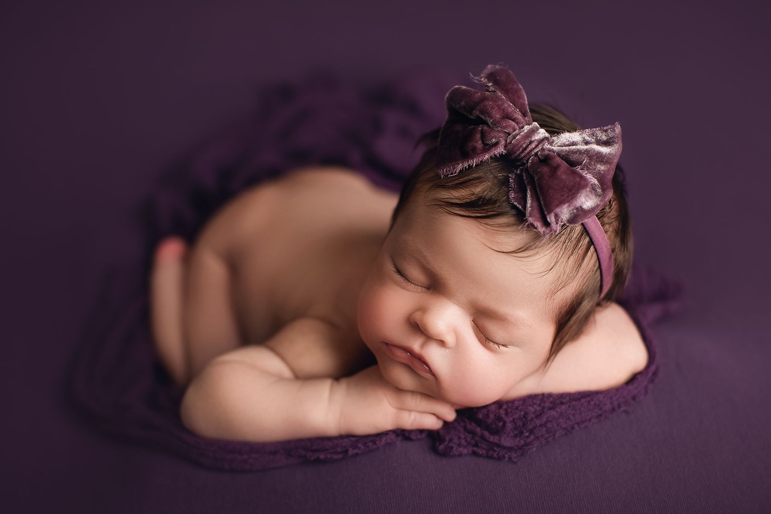 Newborn baby sleeping on purple fabric wearing delicate bow headpiece. Dubai Photographer