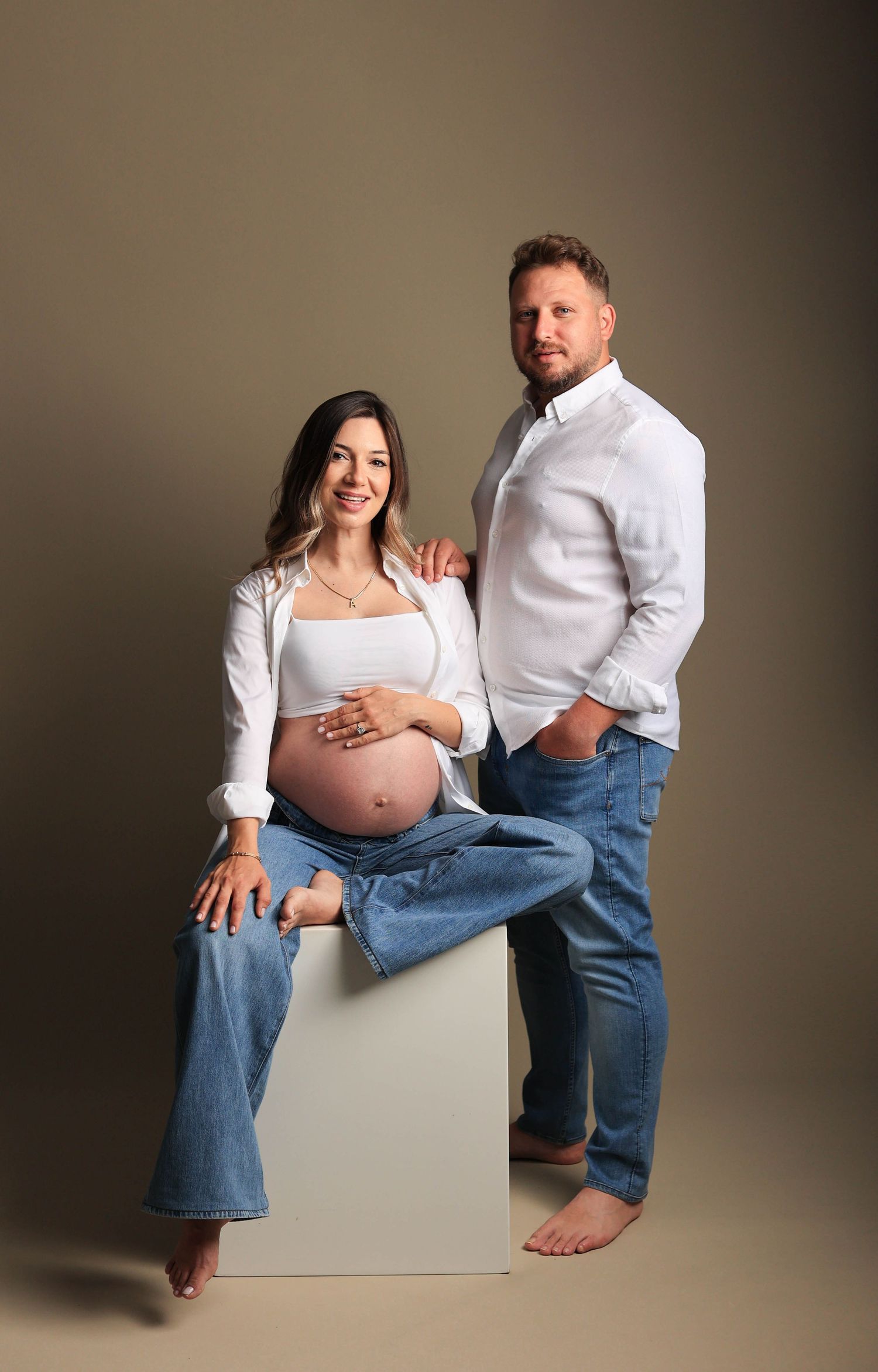 Casual maternity portrait featuring couple in coordinated white tops and denim on simple beige backdrop.Dubai Photographer