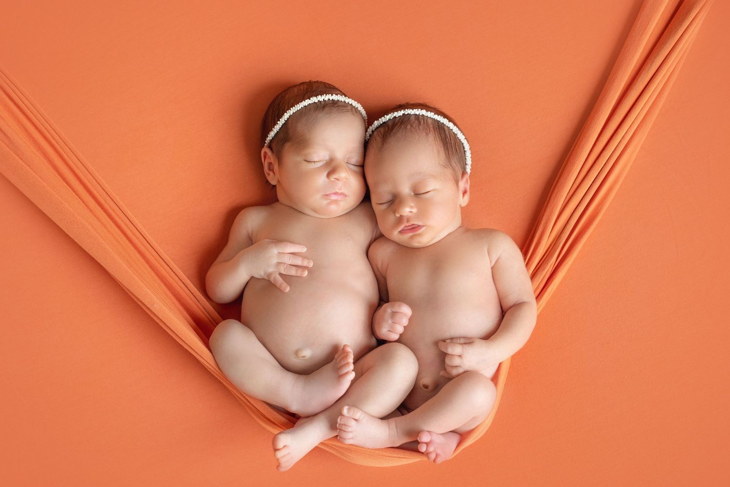 Two sleeping newborns wearing headbands on coral-colored fabric backdrop. Dubai Photographer