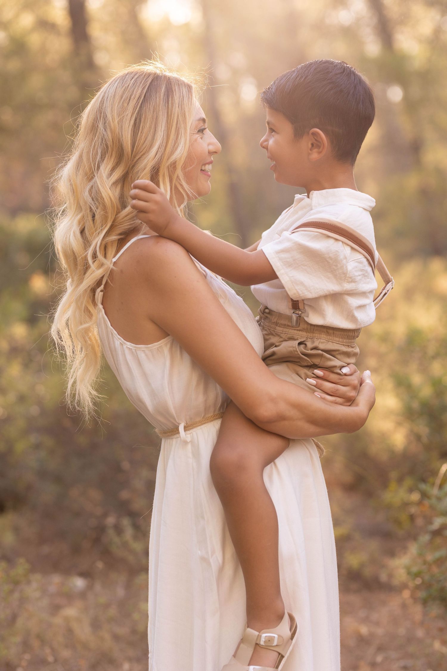 Mother and child sharing a tender moment in golden sunlit forest setting while wearing white summer attire. Dubai Photographer