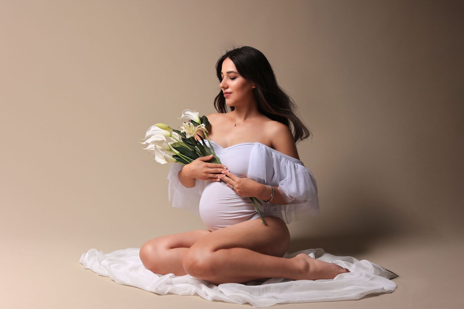 Expectant woman in white dress holds calla lilies while sitting on floor. Dubai Photographer