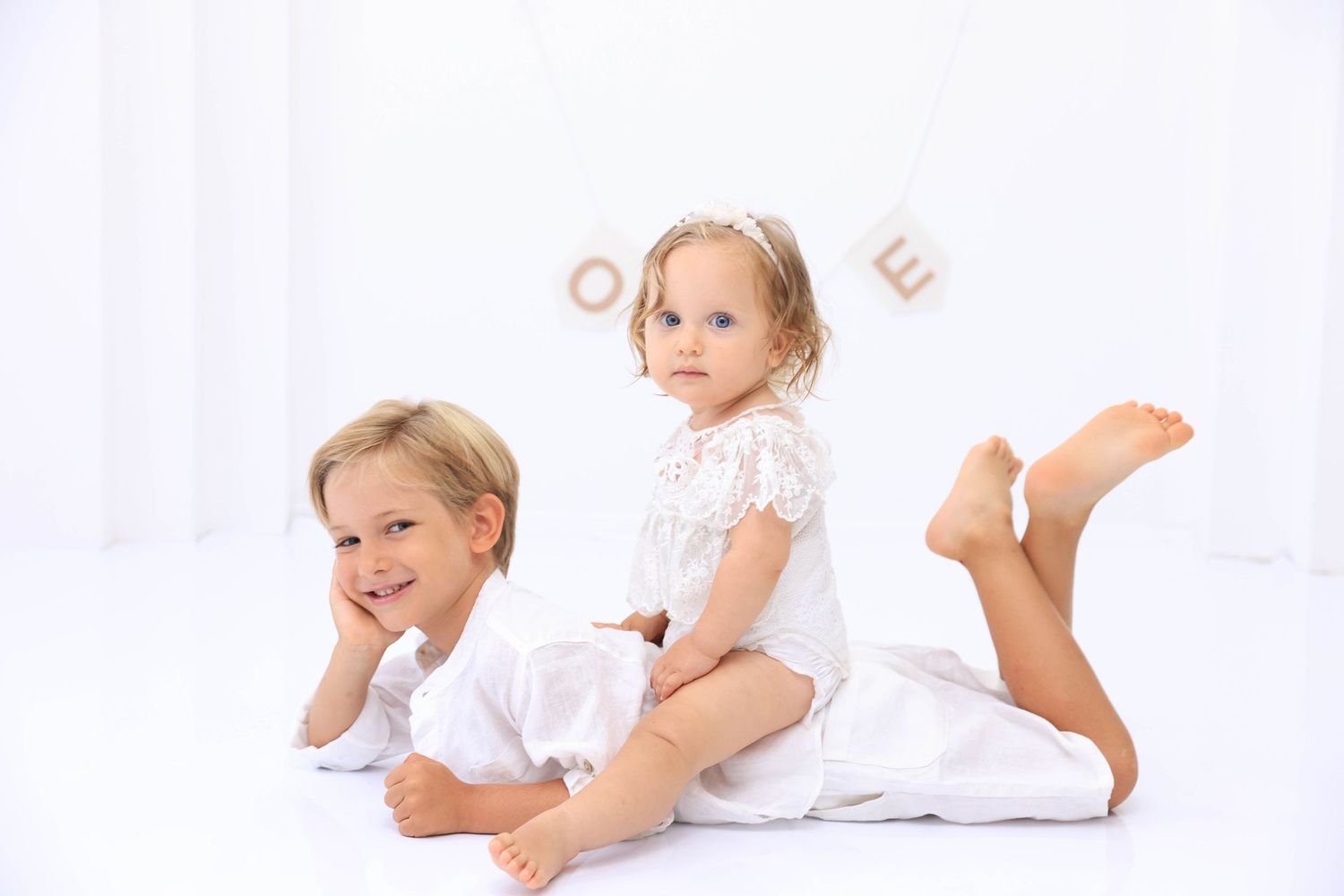 Four young children wearing white clothing lay on a light background, playing and smiling together. Dubai Photographer