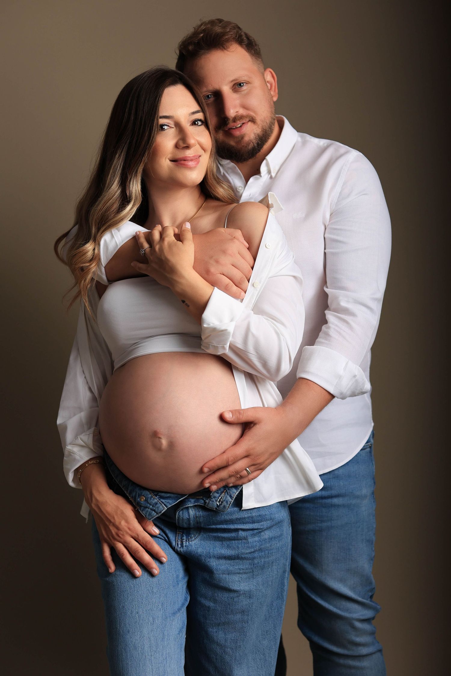 Intimate maternity photo of expecting couple in white shirts embracing against neutral background. Dubai Photographer