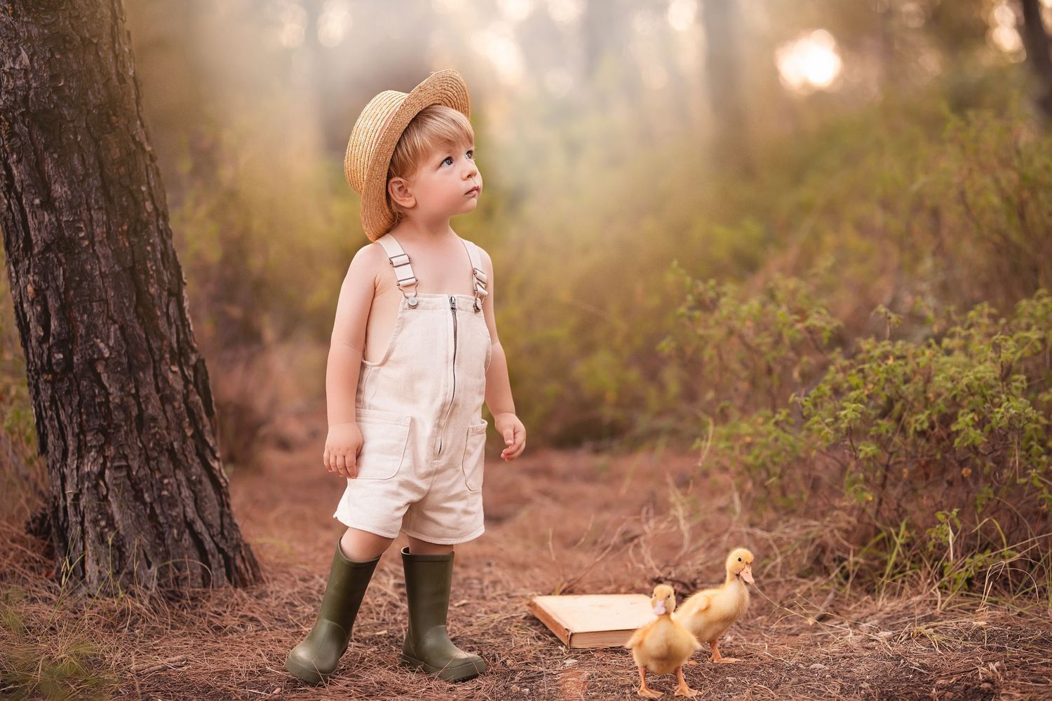 Child in overalls and straw hat standing with baby duck on woodland path at sunset. Dubai Photographer