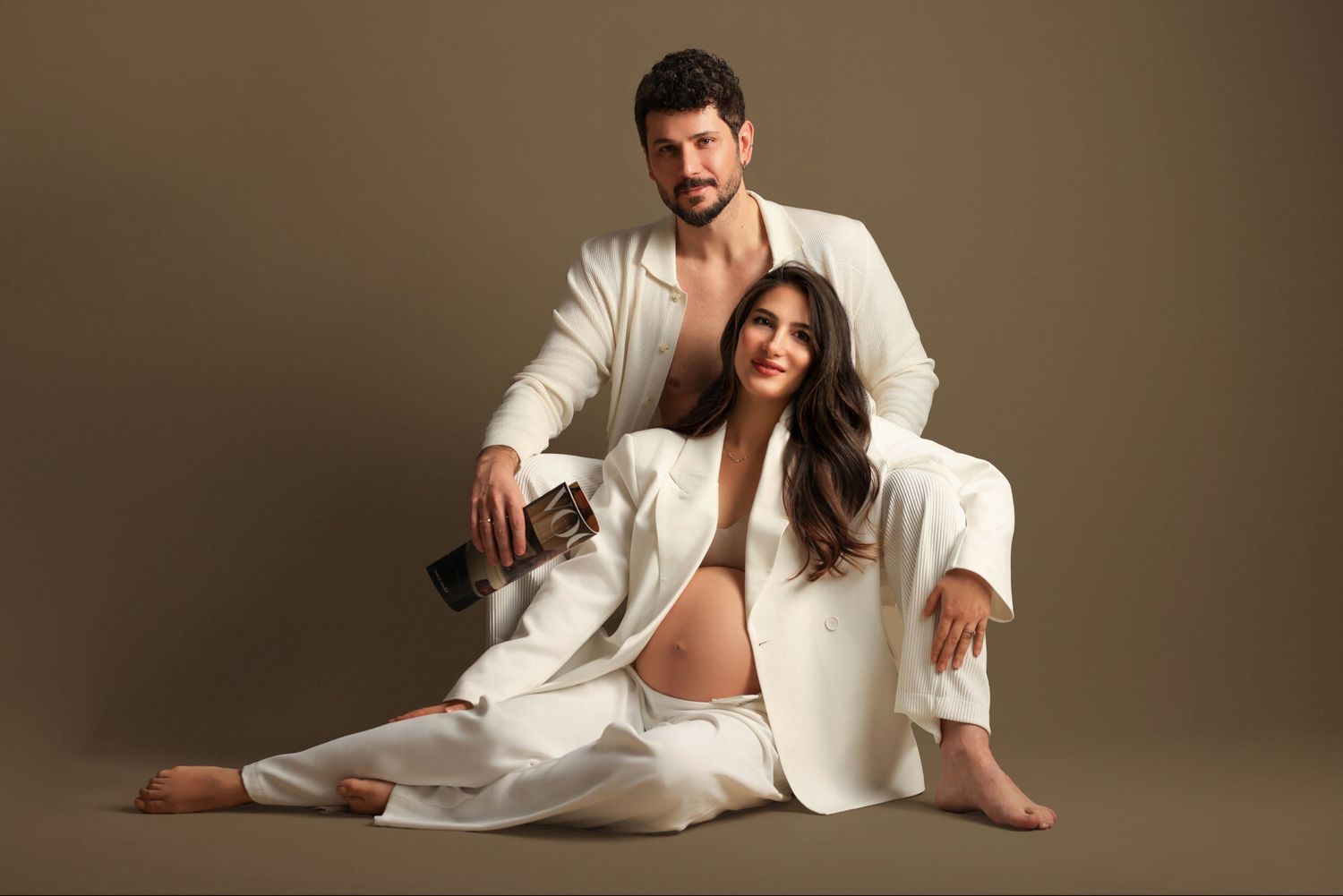 Artistic maternity studio portrait with couple in matching white outfits against brown background. Dubai Photographer