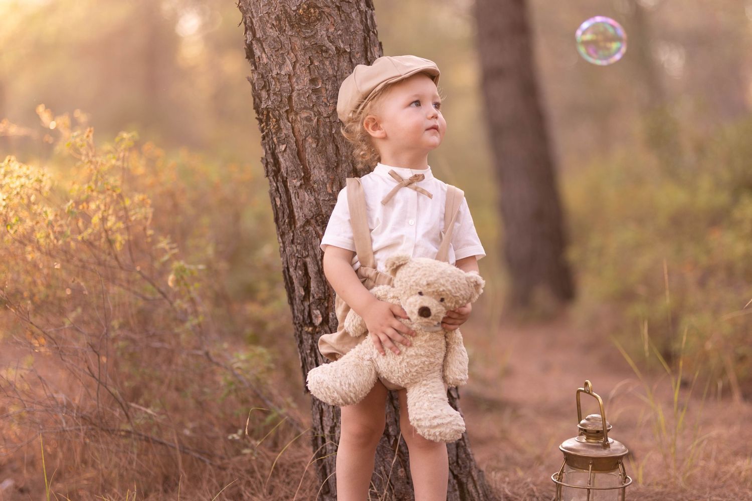 Child in vintage attire holding teddy bear with bubble floating nearby in forest setting. Dubai Photographer