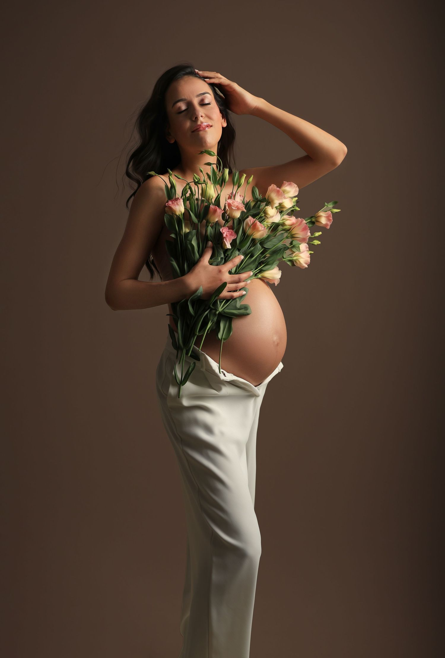 A pregnant woman in a white dress holds a bouquet of pink flowers in a moody maternity portrait against brown backdrop. Dubai Photographer
