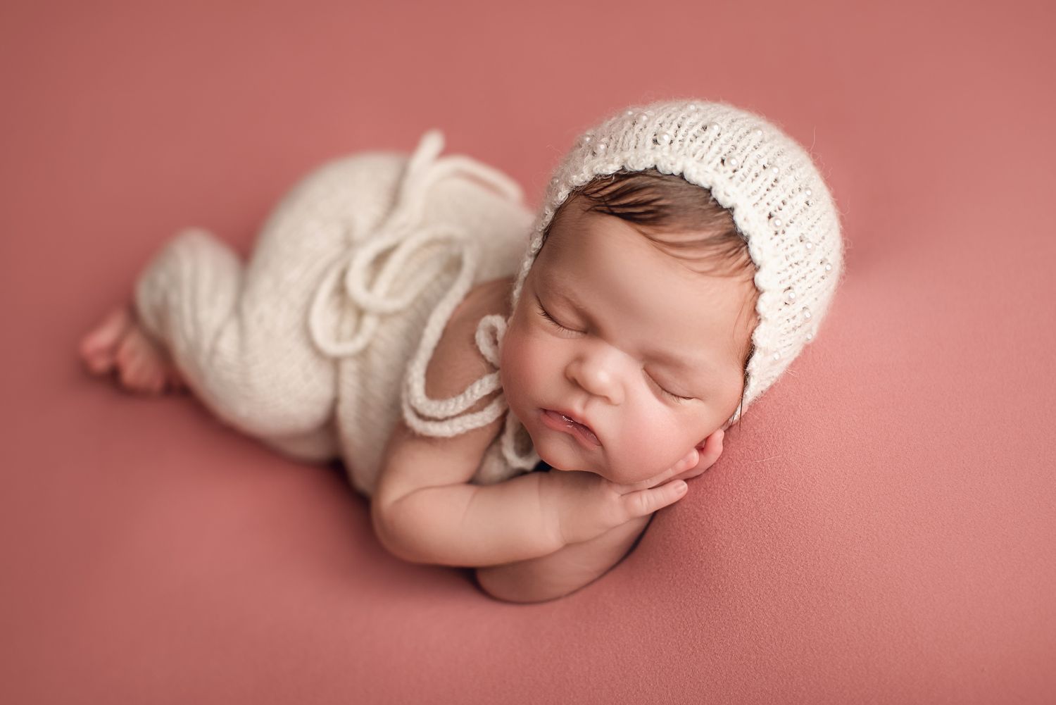 Sleeping newborn wearing white knit bonnet posed on pink backdrop. Dubai Photographer