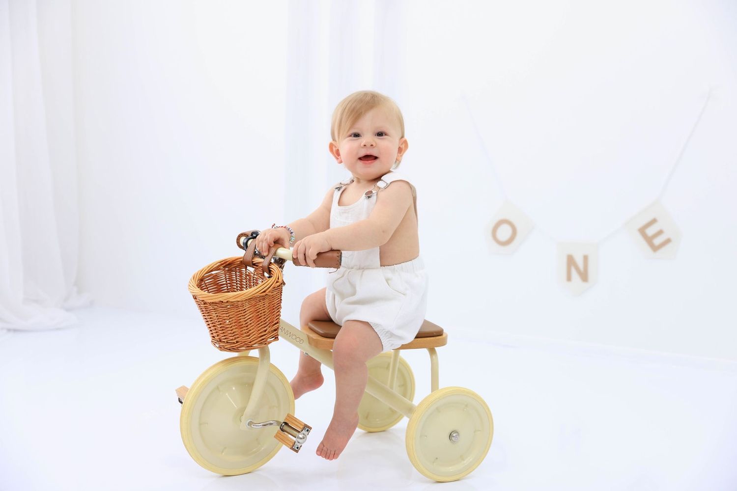 A happy baby in white sits on a vintage-style tricycle with a wicker basket against a light backdrop with letter decorations. Dubai Photogra