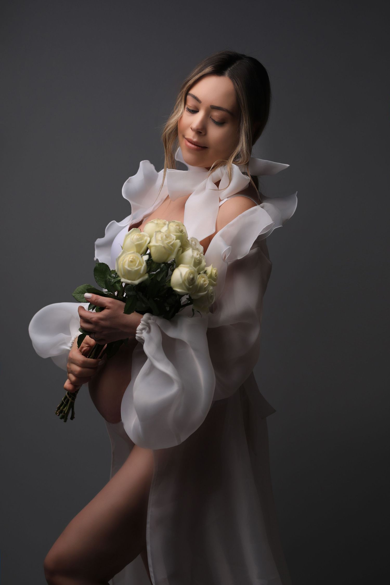 Artistic photo of person in white ruffled dress holding cream roses against dark background. Dubai Photographer