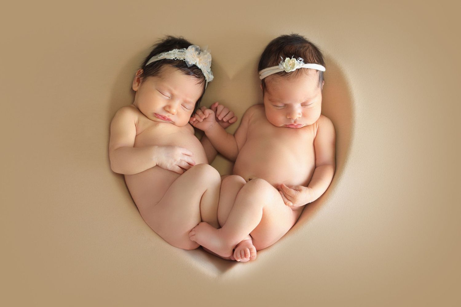 Two sleeping newborn babies nestled together in a heart-shaped frame on a beige background. Dubai Photographer