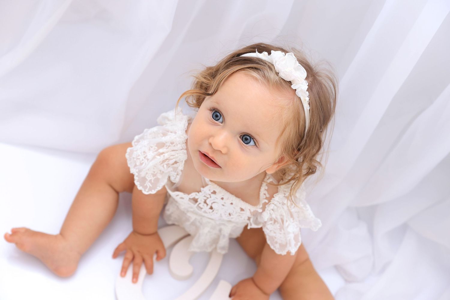 Baby wearing delicate white lace dress and headband sits on soft white tulle fabric. Dubai Photographer