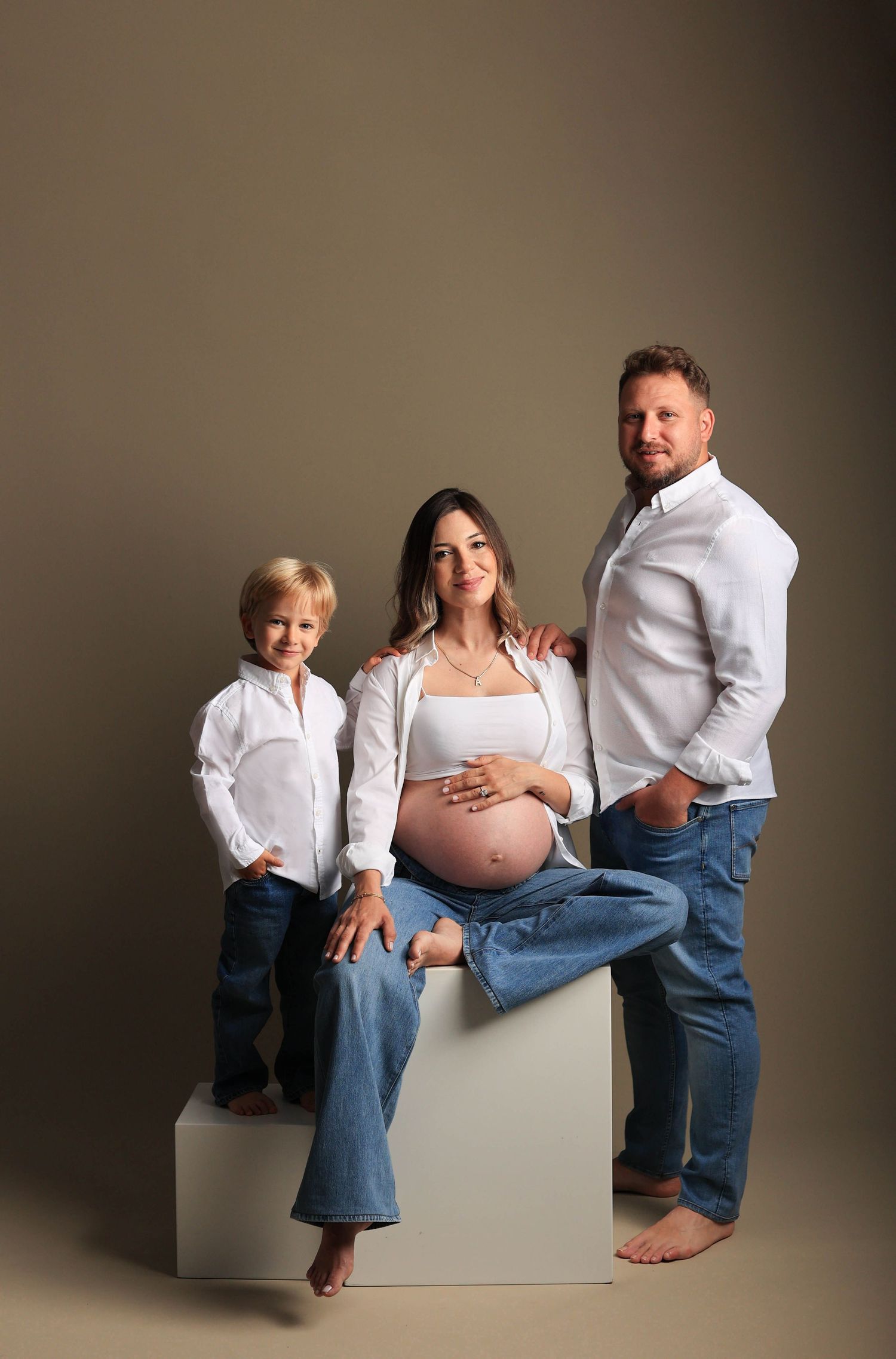 A family maternity photo with everyone wearing white and blue posing together on a white cube against a neutral backdrop.