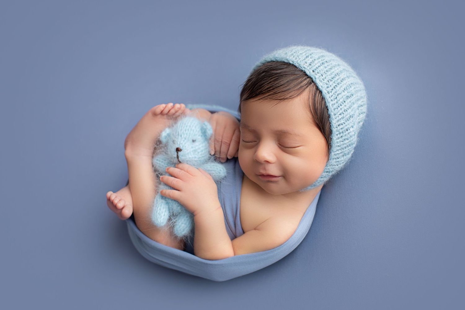 Sleeping newborn in light blue bonnet holding a tiny teddy bear in blue bowl. Dubai Photographer
