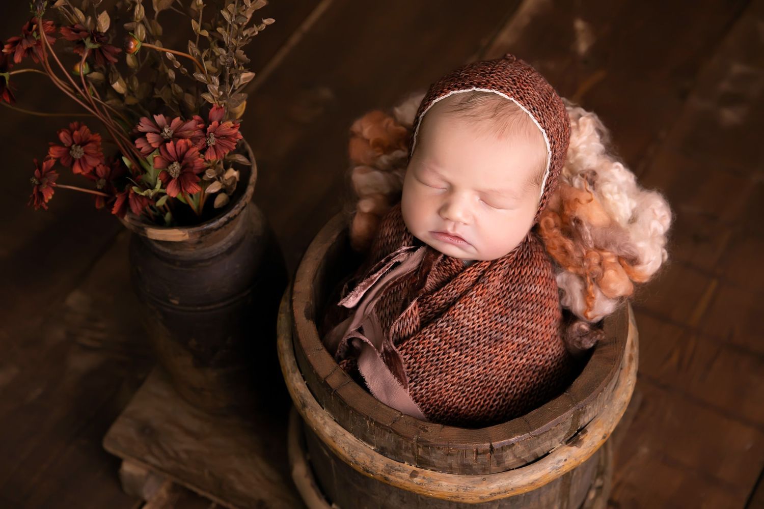 Sleeping newborn in knit outfit posed in rustic wooden barrel with floral decor. Dubai Photographer