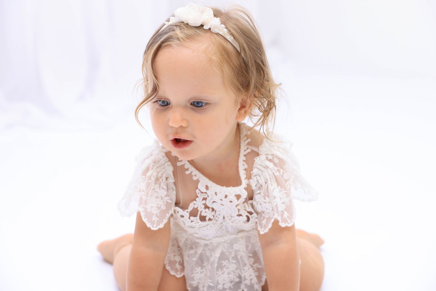 A small child wearing a delicate white lace dress and hair bow sits against a light background. Dubai Photographer
