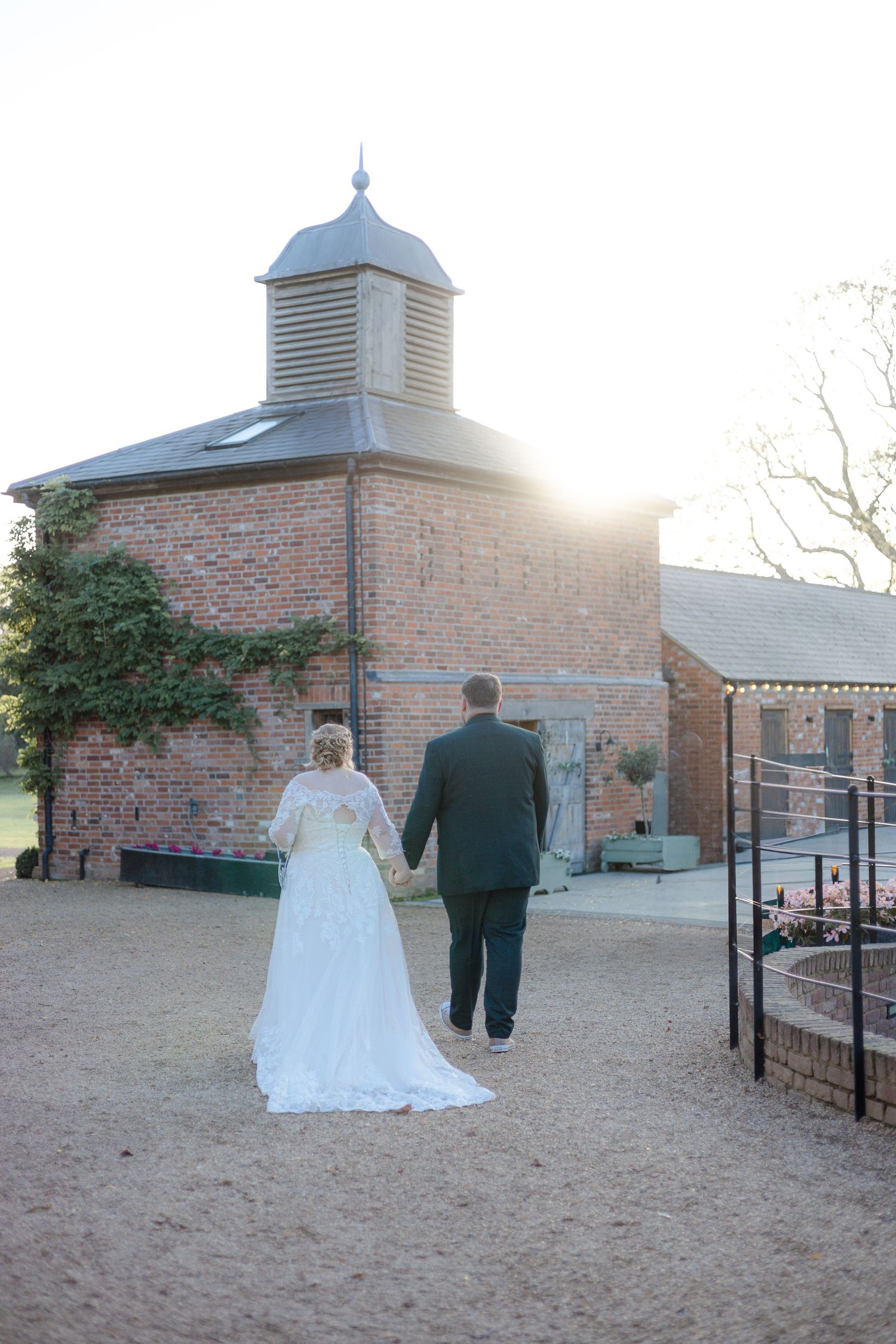 A wedding couple walks along a brick courtyard with architectural features and sunlight.