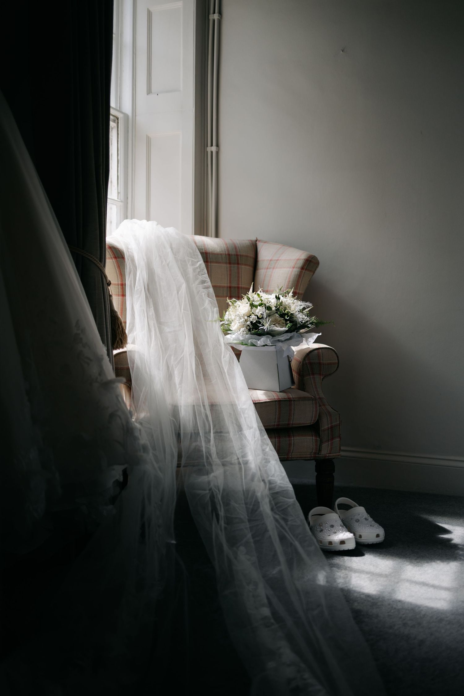 Ethereal bridal gown and veil detail by window in moody natural lighting.