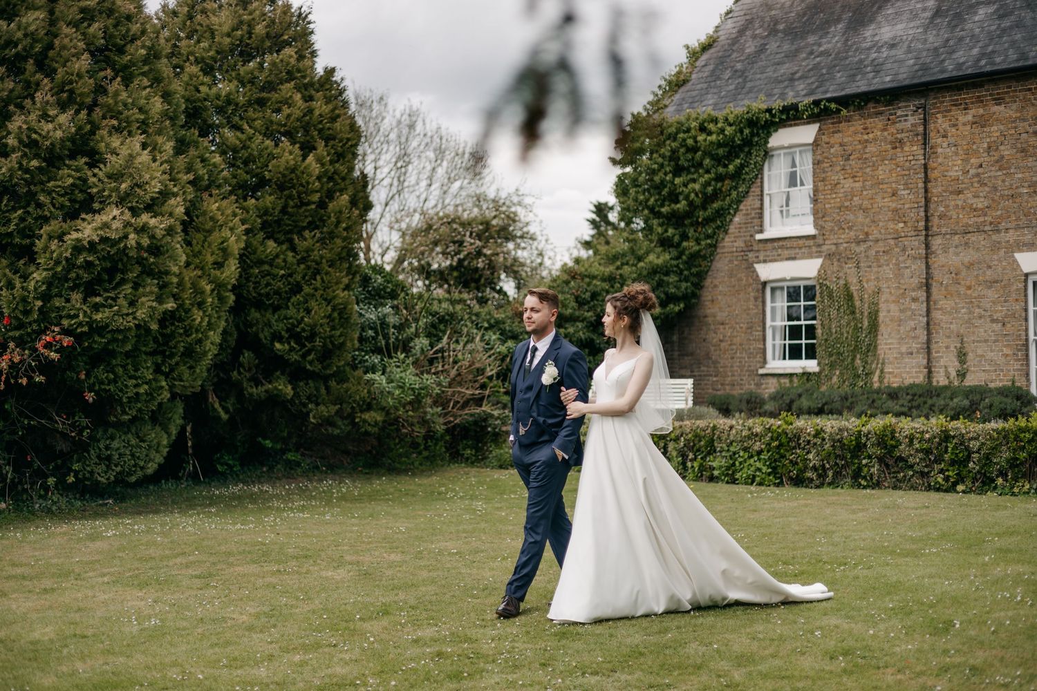Wedding couple poses in front of a traditional stone house with manicured gardens.