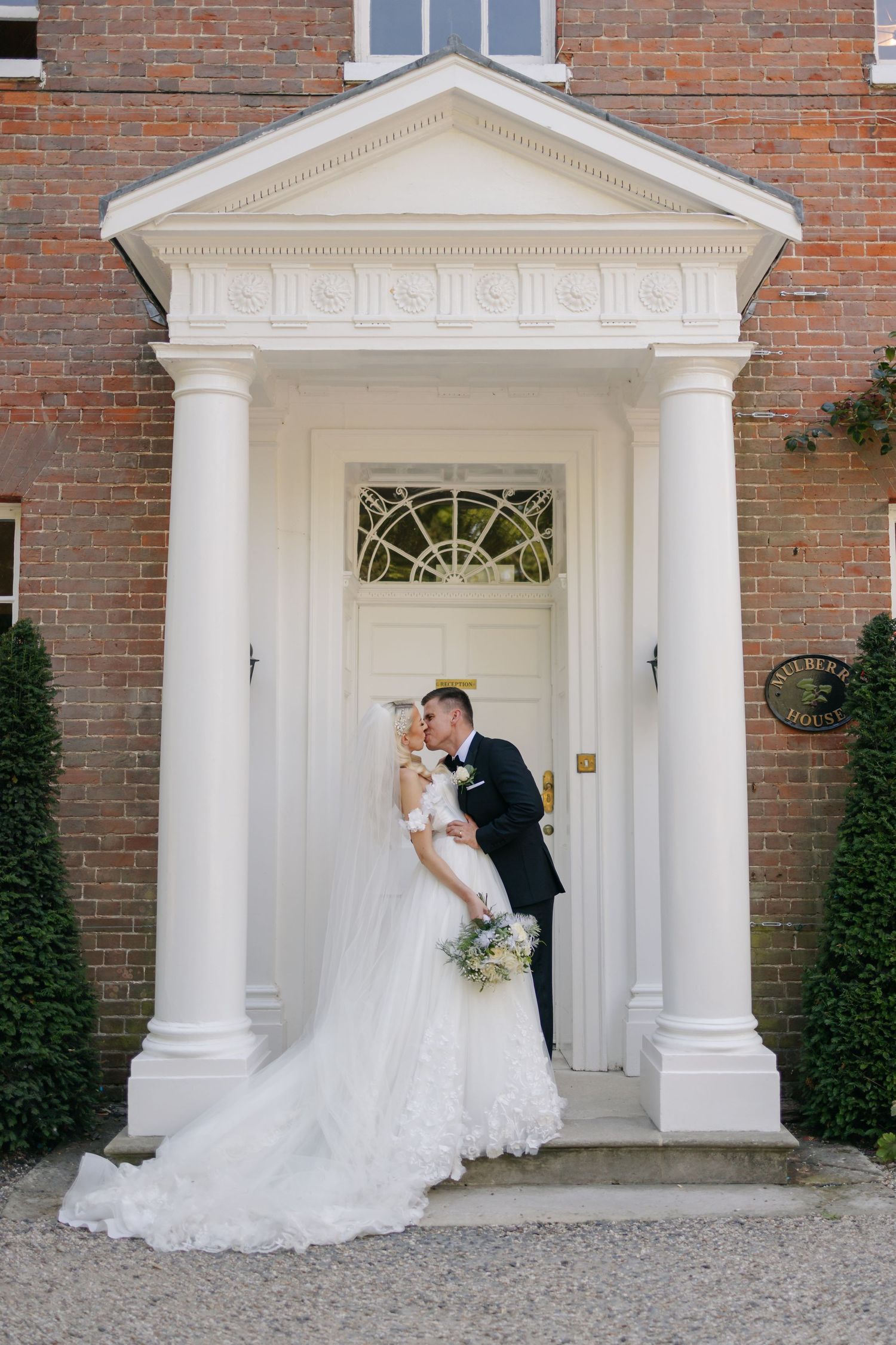 Wedding couple poses in front of a classic brick building with white columned entrance.