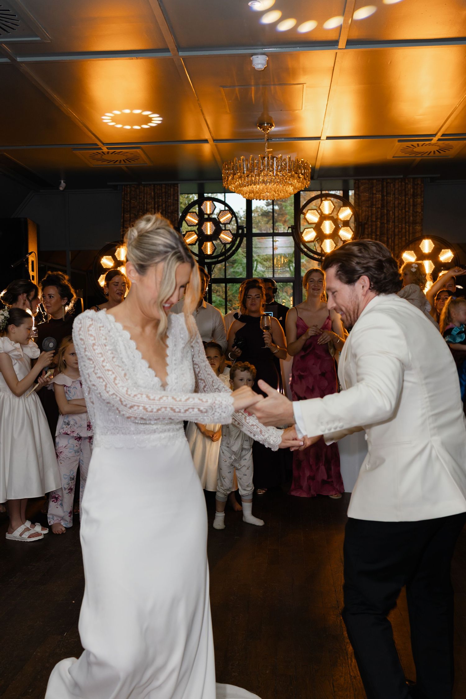 Bride in lace wedding gown and groom in white jacket share an energetic first dance surrounded by cheering guests.