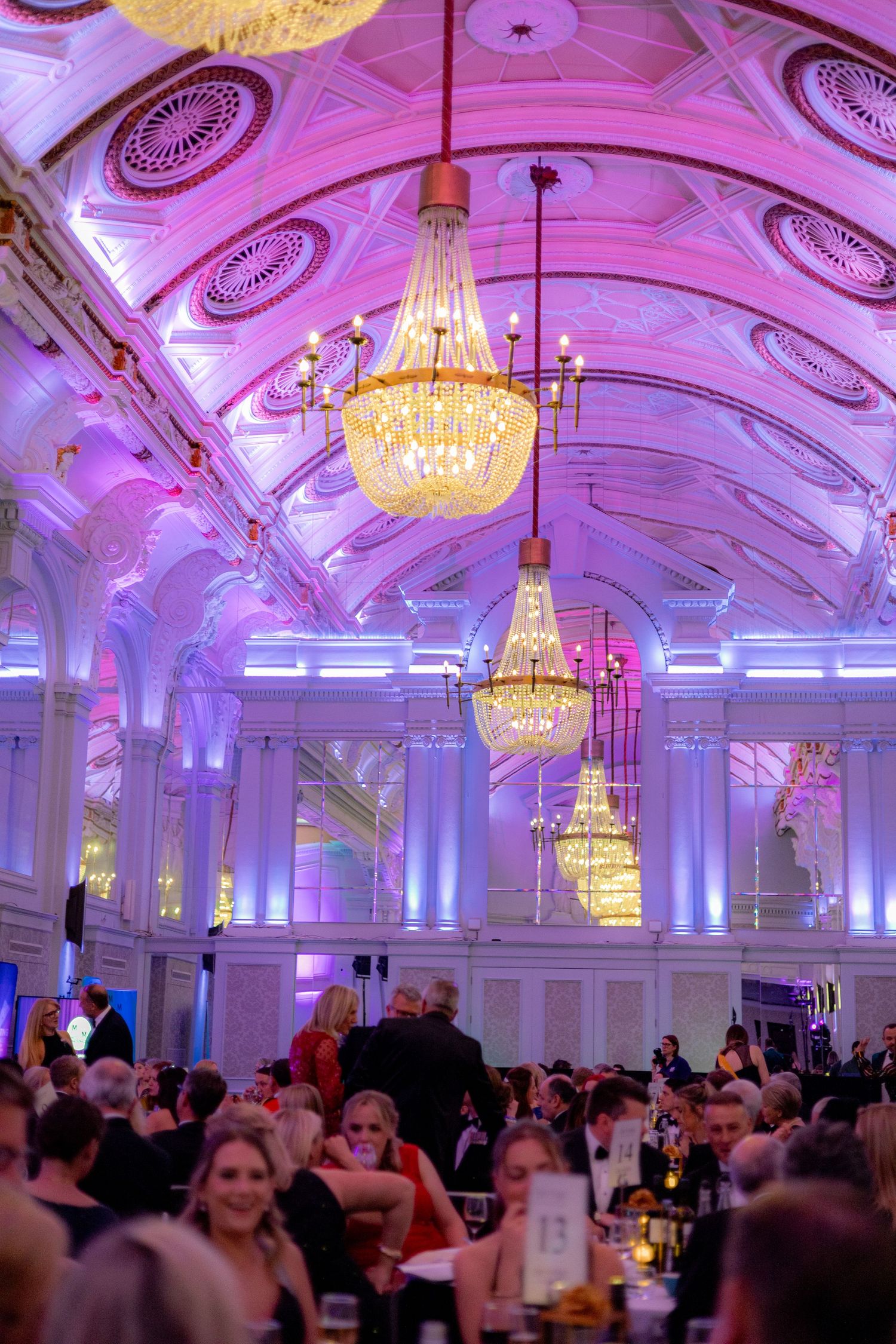 Elegant ballroom with ornate chandeliers and purple lighting during a formal event.