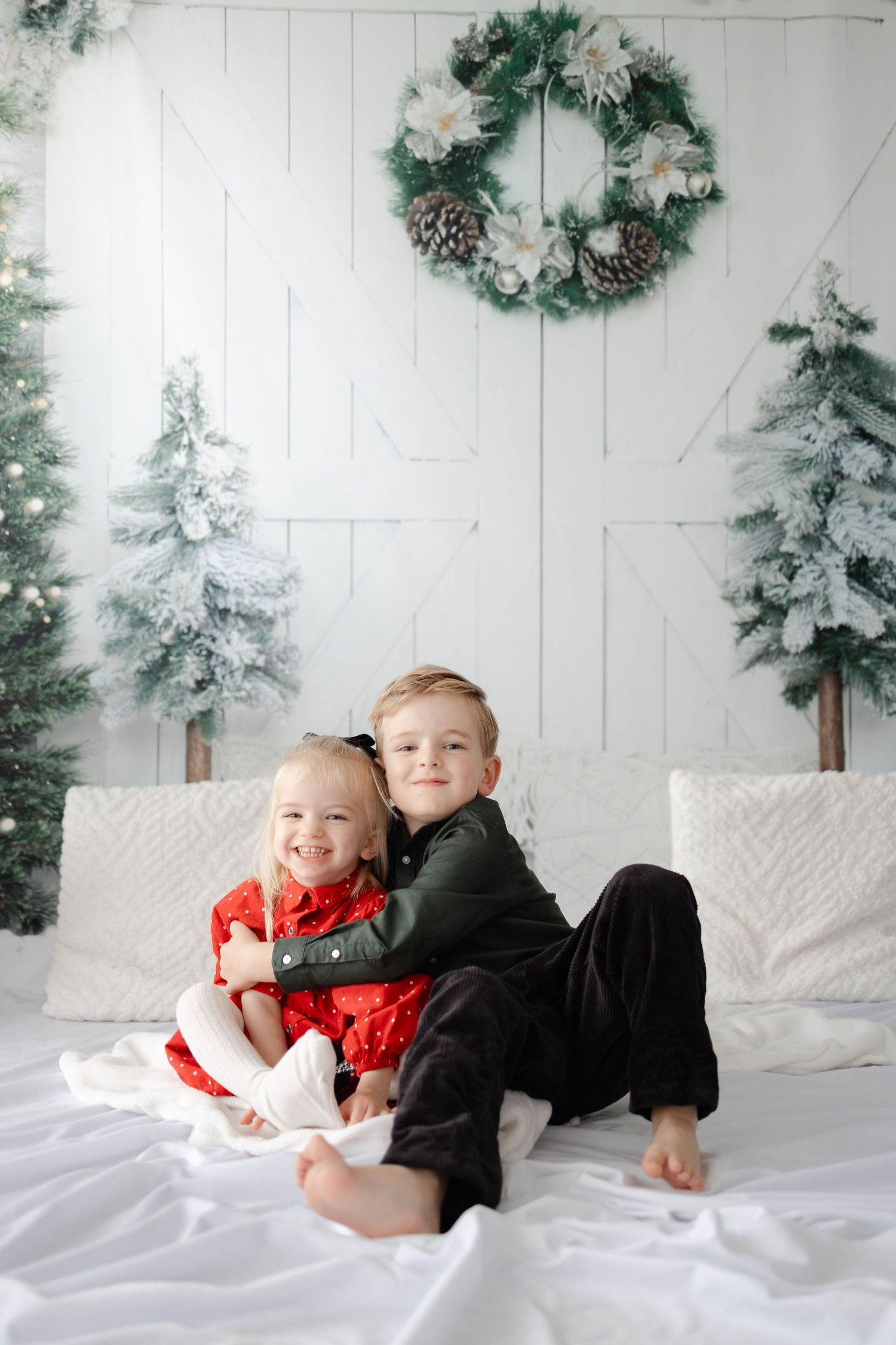Children in coordinating outfits playing on white bedding with winter decorations and flocked trees.