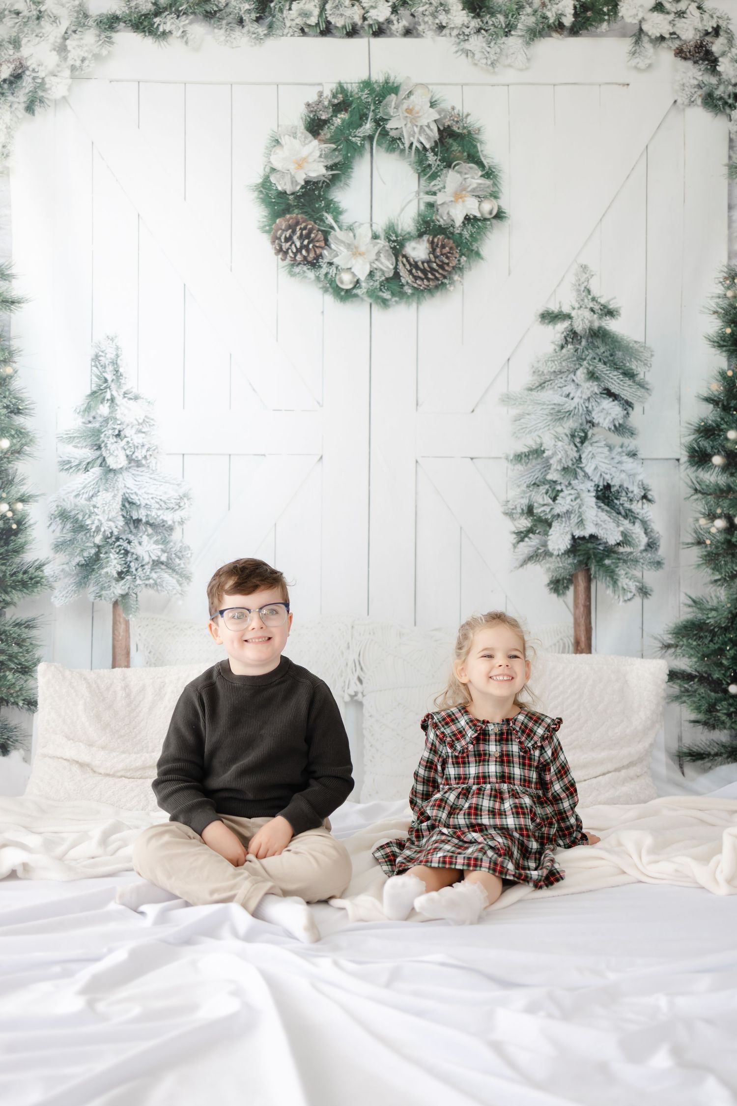 A cozy Christmas portrait setup with two children sitting on white bedding surrounded by snowy trees and a wreath.