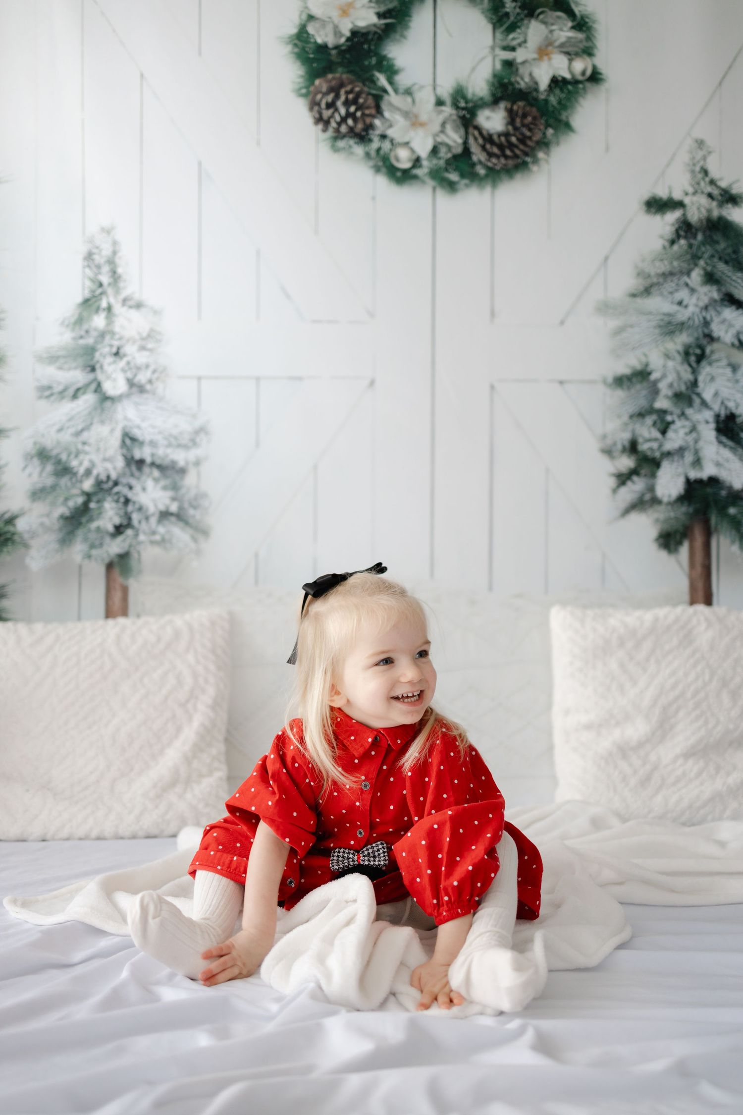 Baby in red polka dot dress crawling on white bedding with winter holiday backdrop.