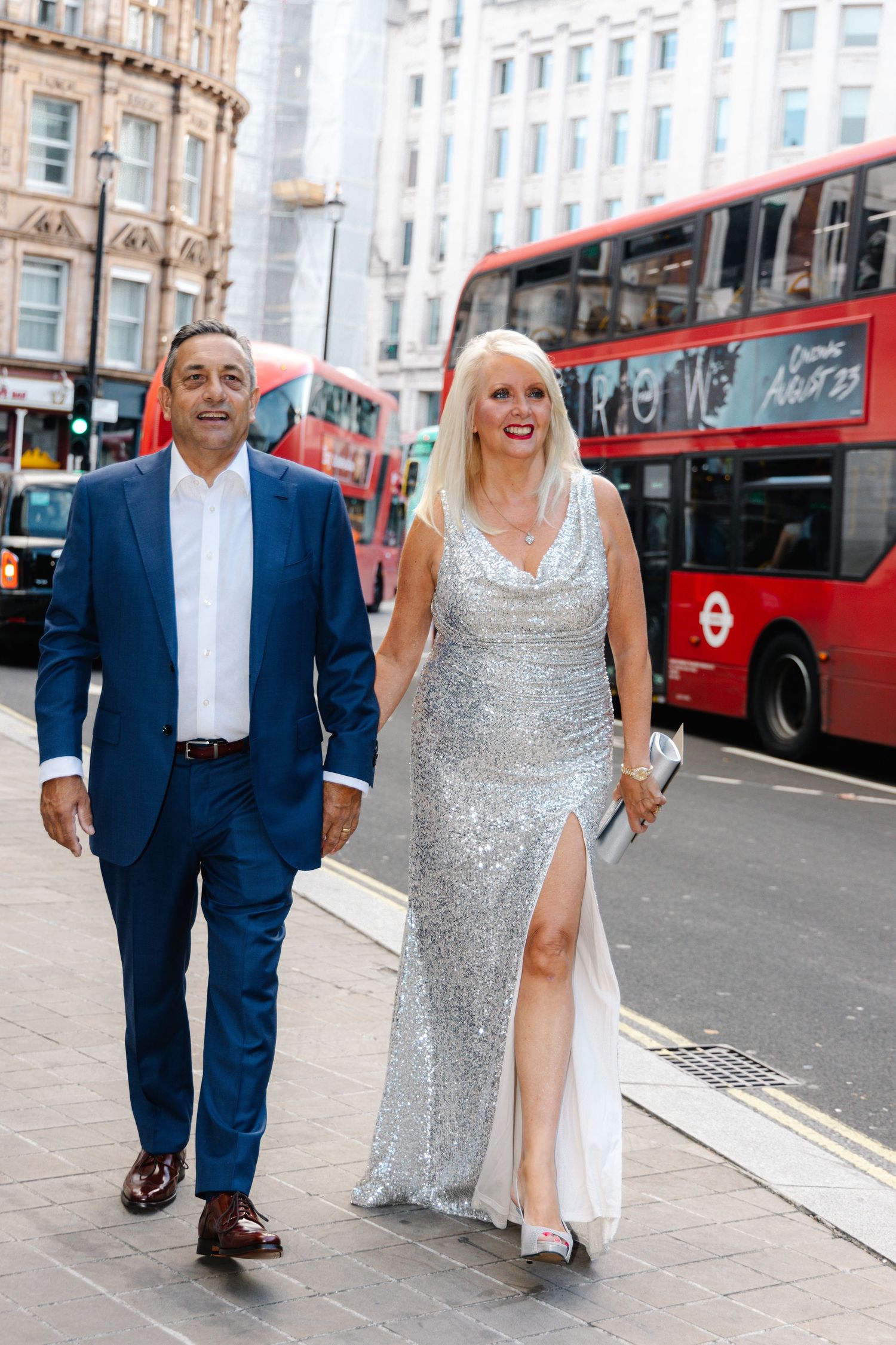 A couple walks down a London street with red buses in the background.