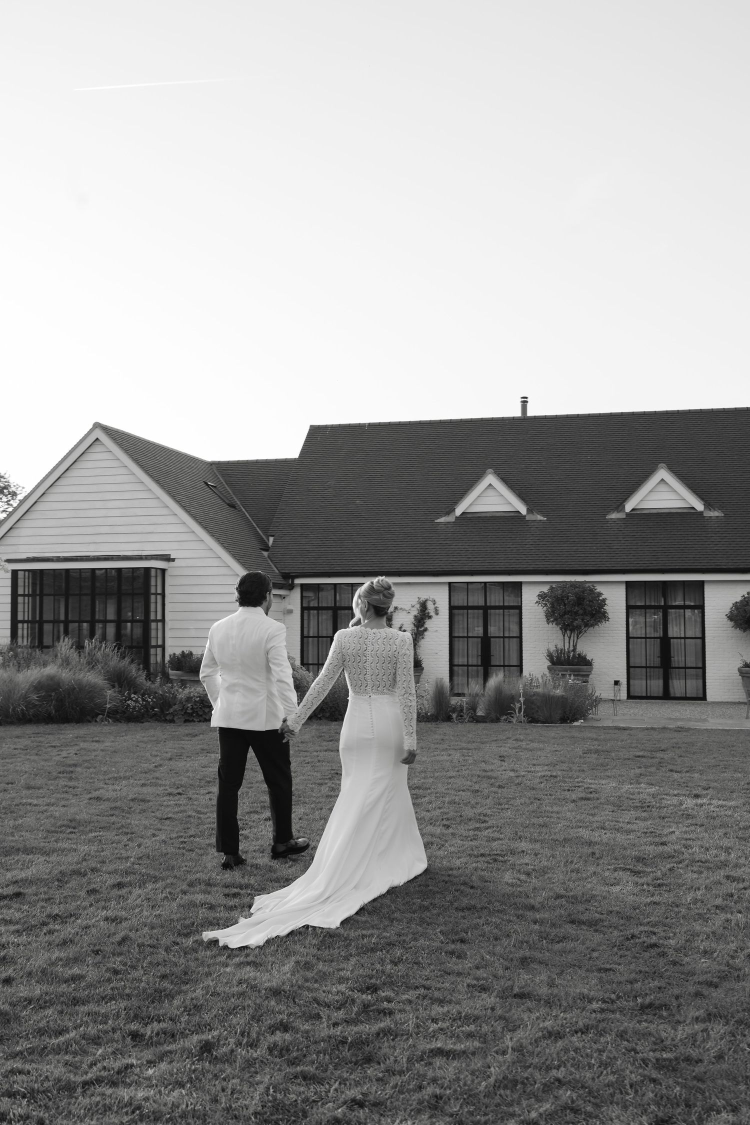 Black and white photo of a bride and groom holding hands walking toward a white farmhouse wedding venue.