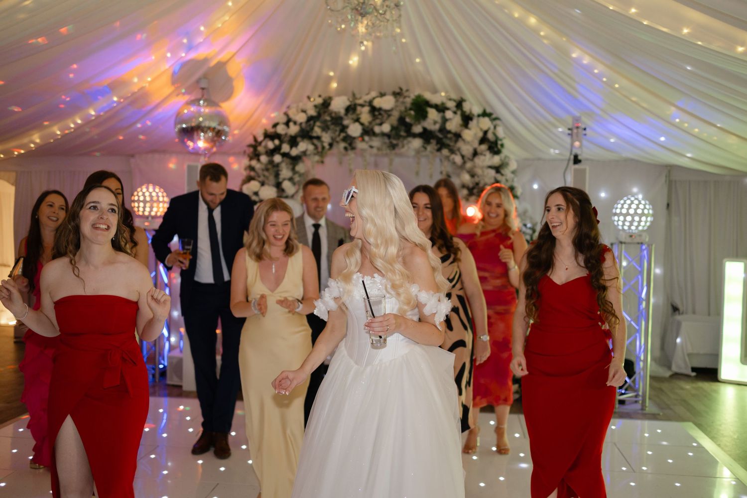 Wedding reception dance floor filled with guests in red dresses under elegant white tent.
