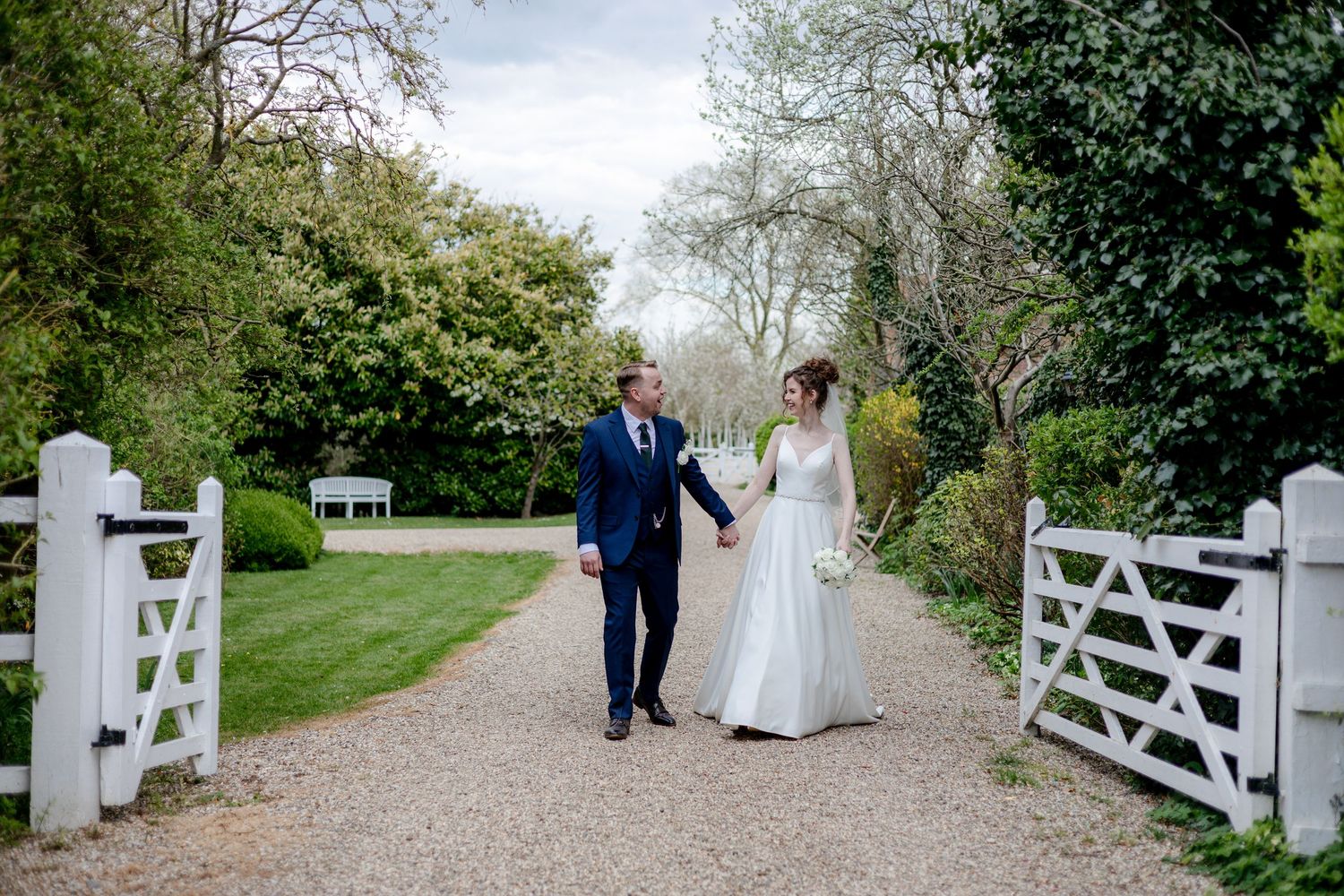 Wedding couple walking along a country path with white garden gates and green foliage.