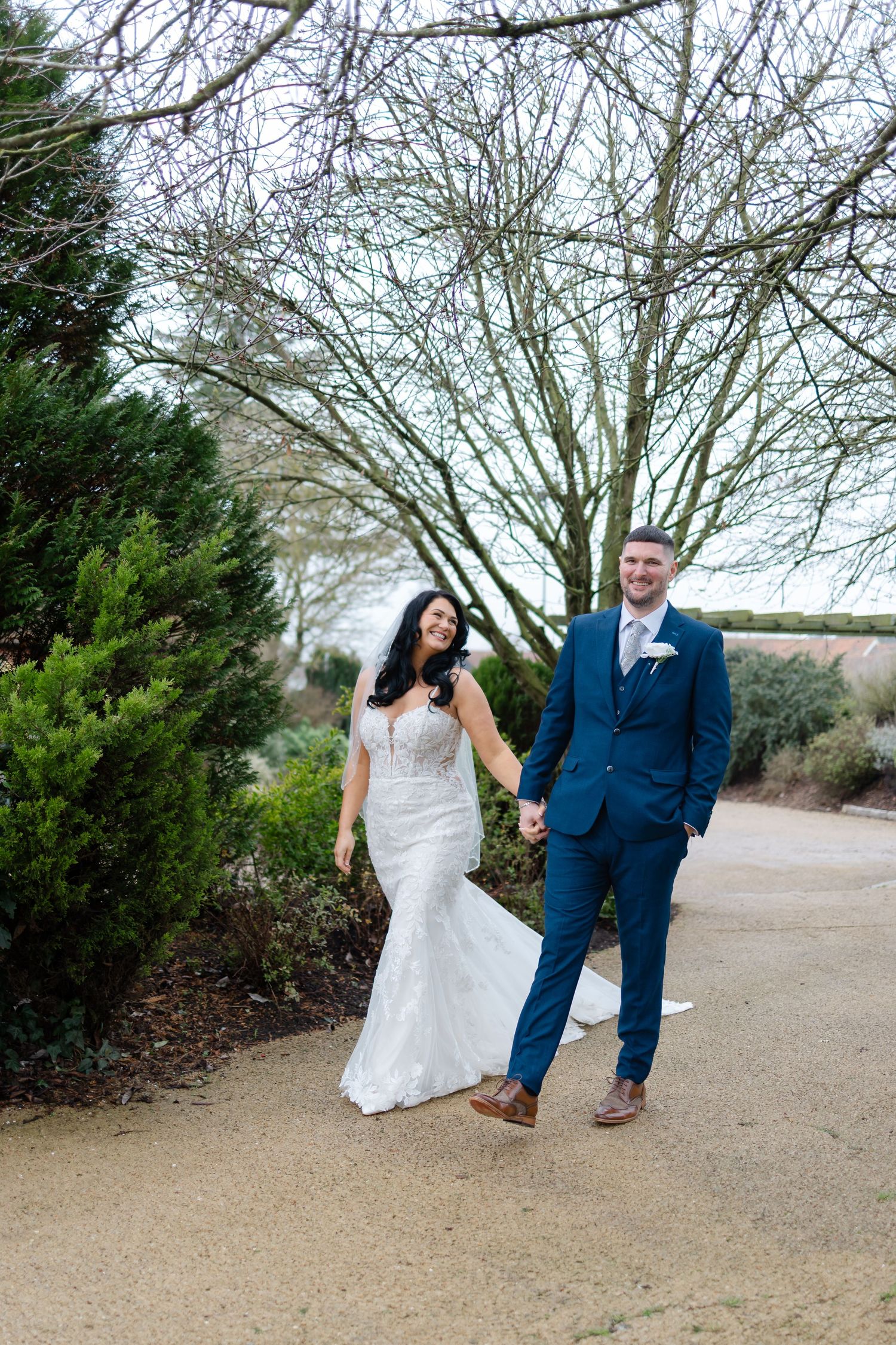 A couple in wedding attire walks along a sandy path surrounded by bare winter trees and greenery.
~ Essex Wedding Photographer