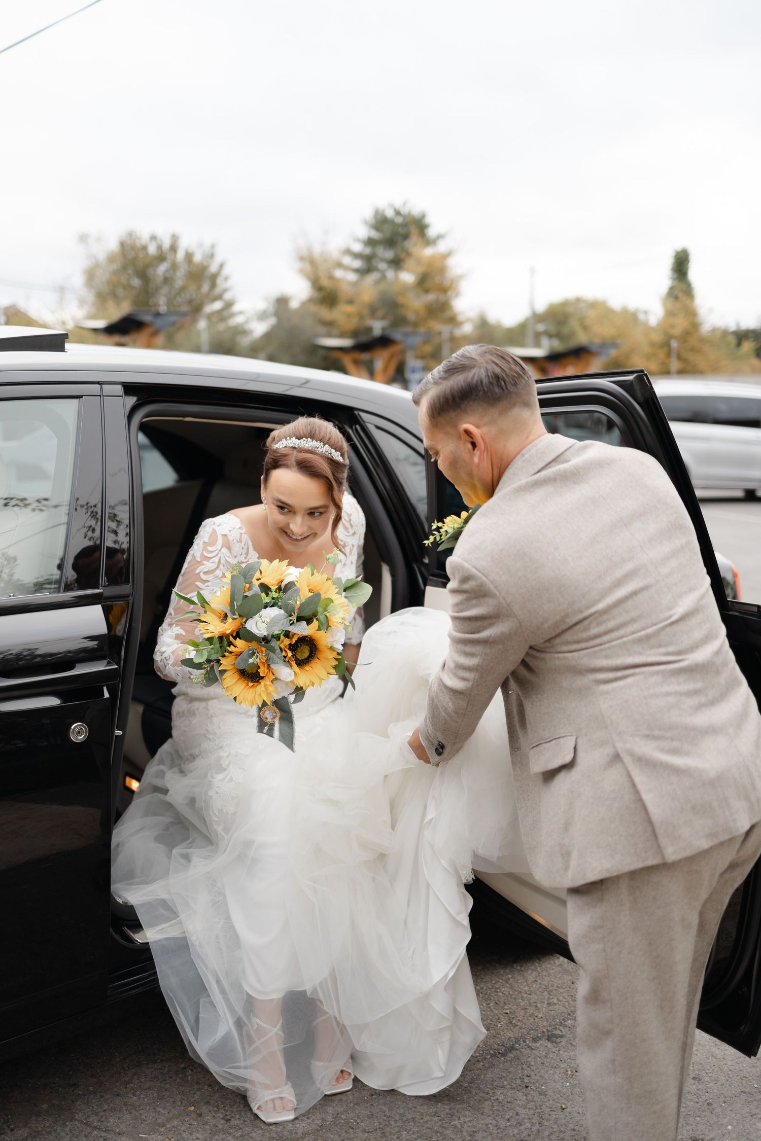 Groom in light gray suit helping bride with sunflower bouquet exit black car.