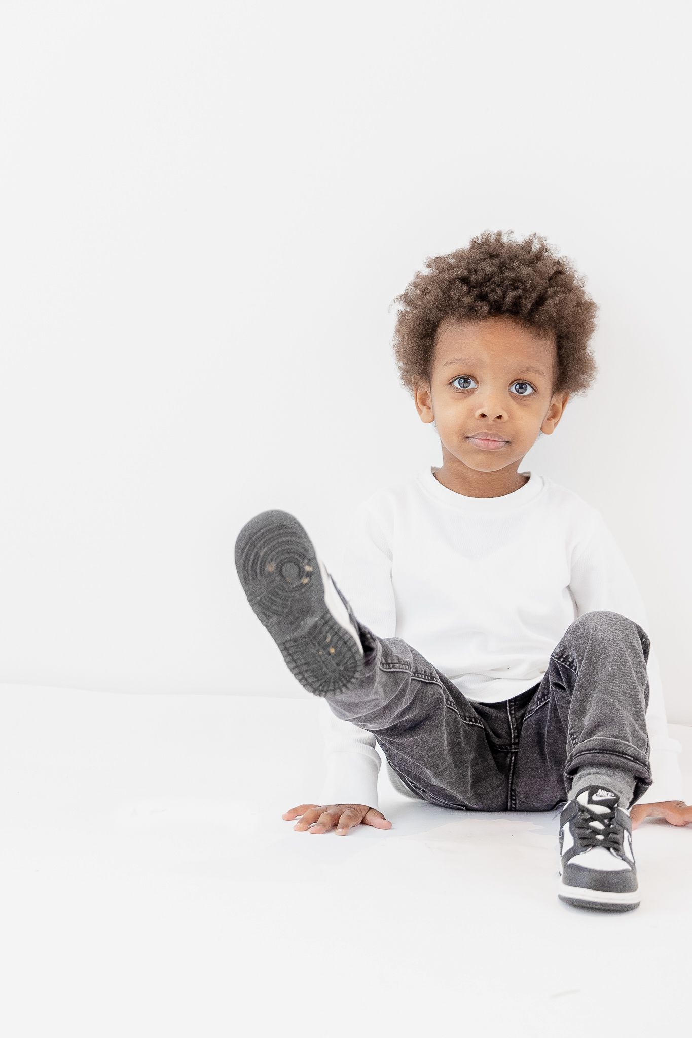 Young child in white shirt and gray jeans sits on white floor showing playful expression.