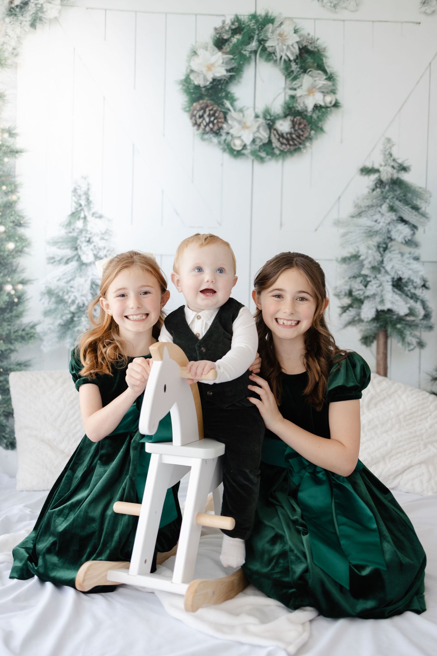 Children in emerald green dresses play with a white rocking horse against a snowy winter backdrop.