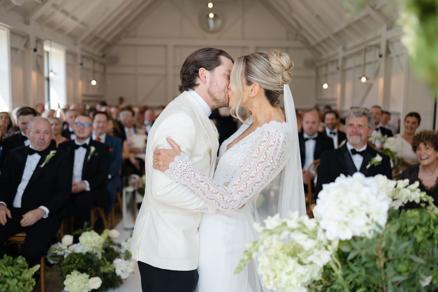 Bride and groom share their first kiss at the altar surrounded by white floral arrangements and wedding guests.