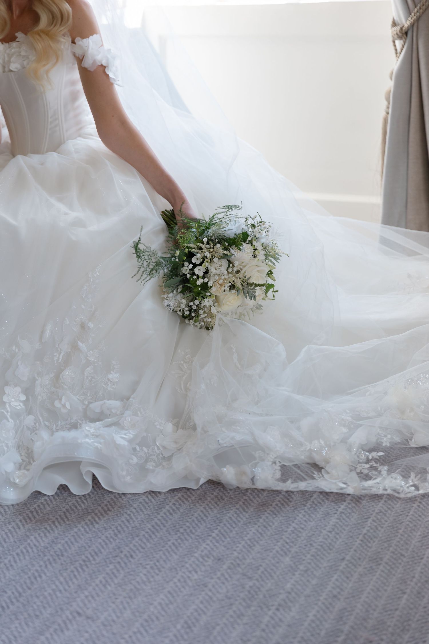 White wedding dress with delicate lace details and a fresh floral bouquet on grey carpet.
