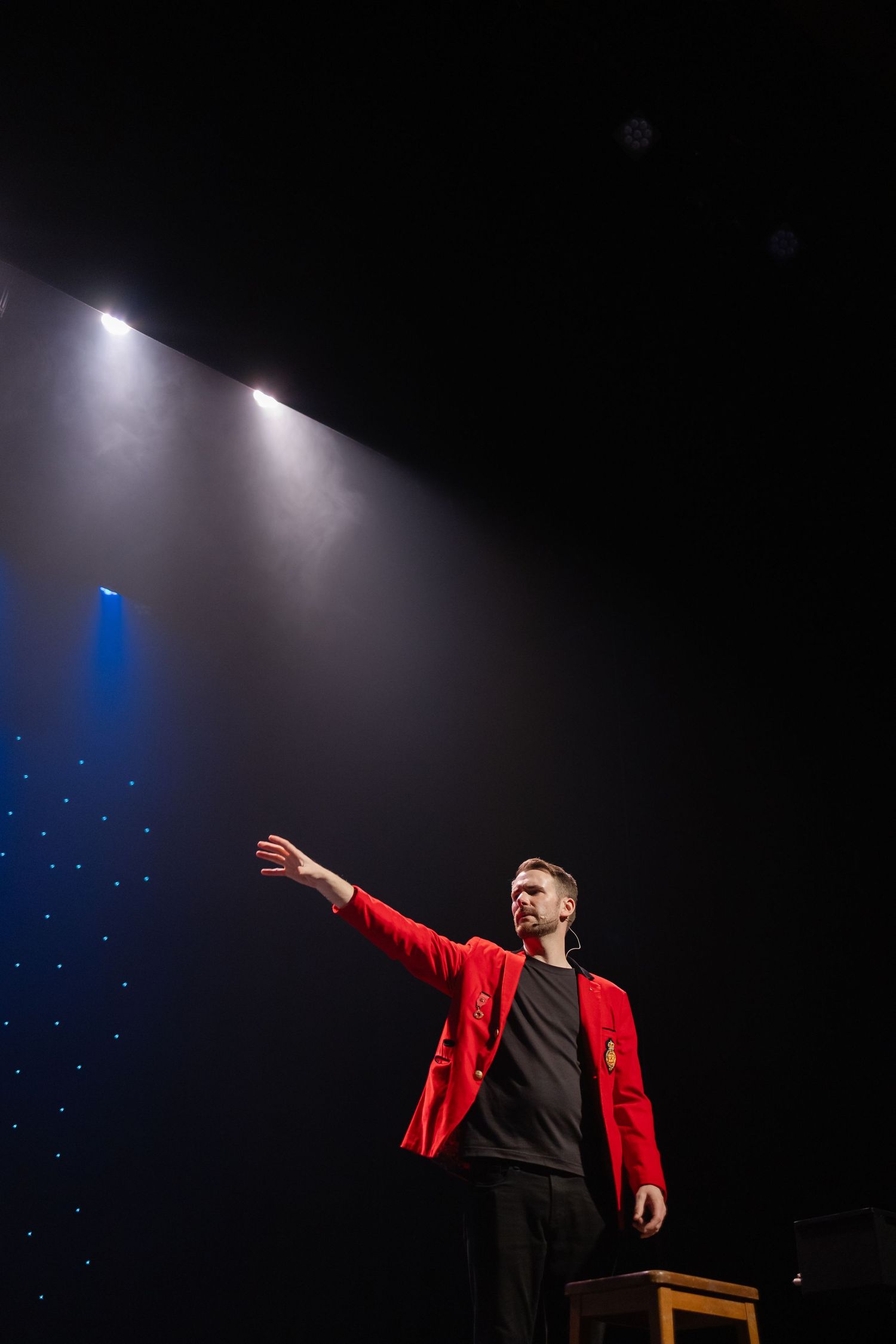Stage performer in red jacket illuminated by dramatic spotlight against dark background.
