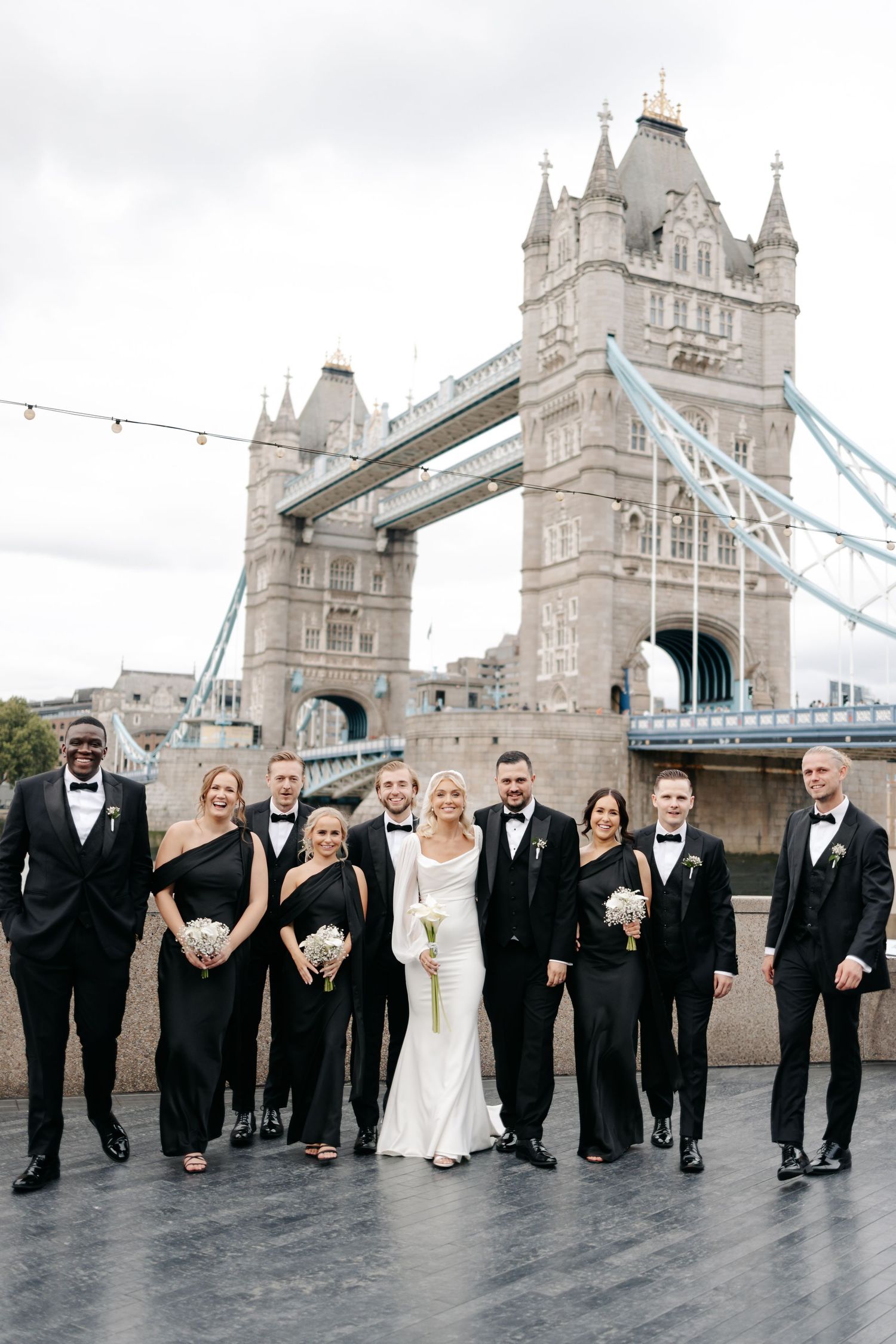 Wedding party in formal attire posing in front of iconic Tower Bridge in London.