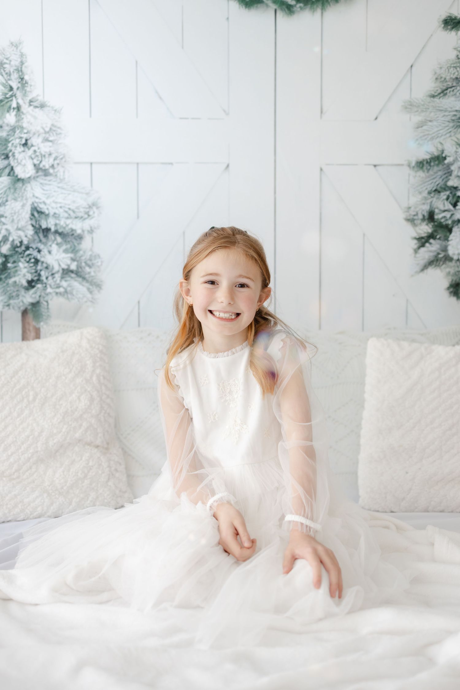Young person in white dress poses on white bedding with winter-themed backdrop and evergreen trees.