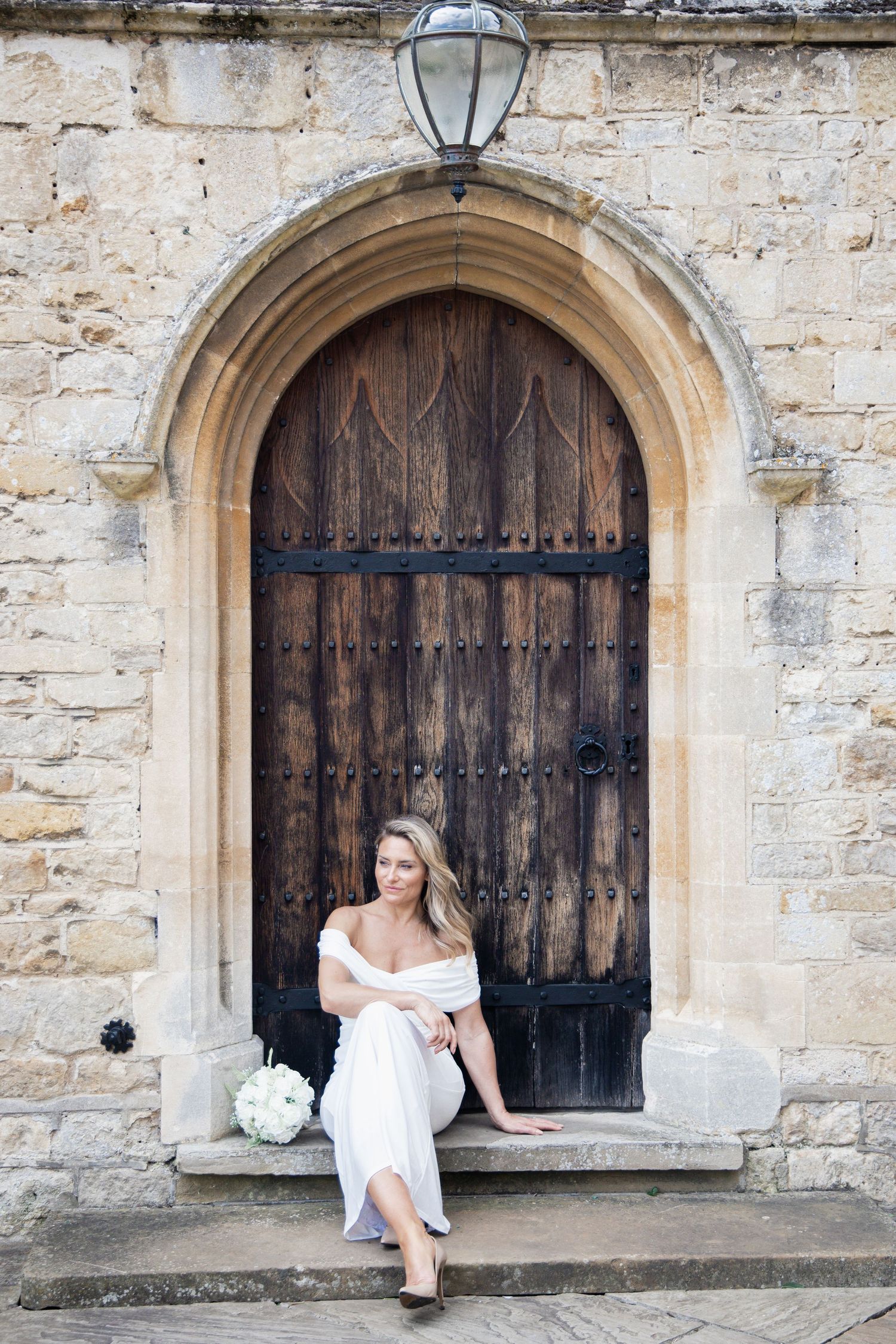 A bride in white sits elegantly in an arched medieval doorway of a stone building.