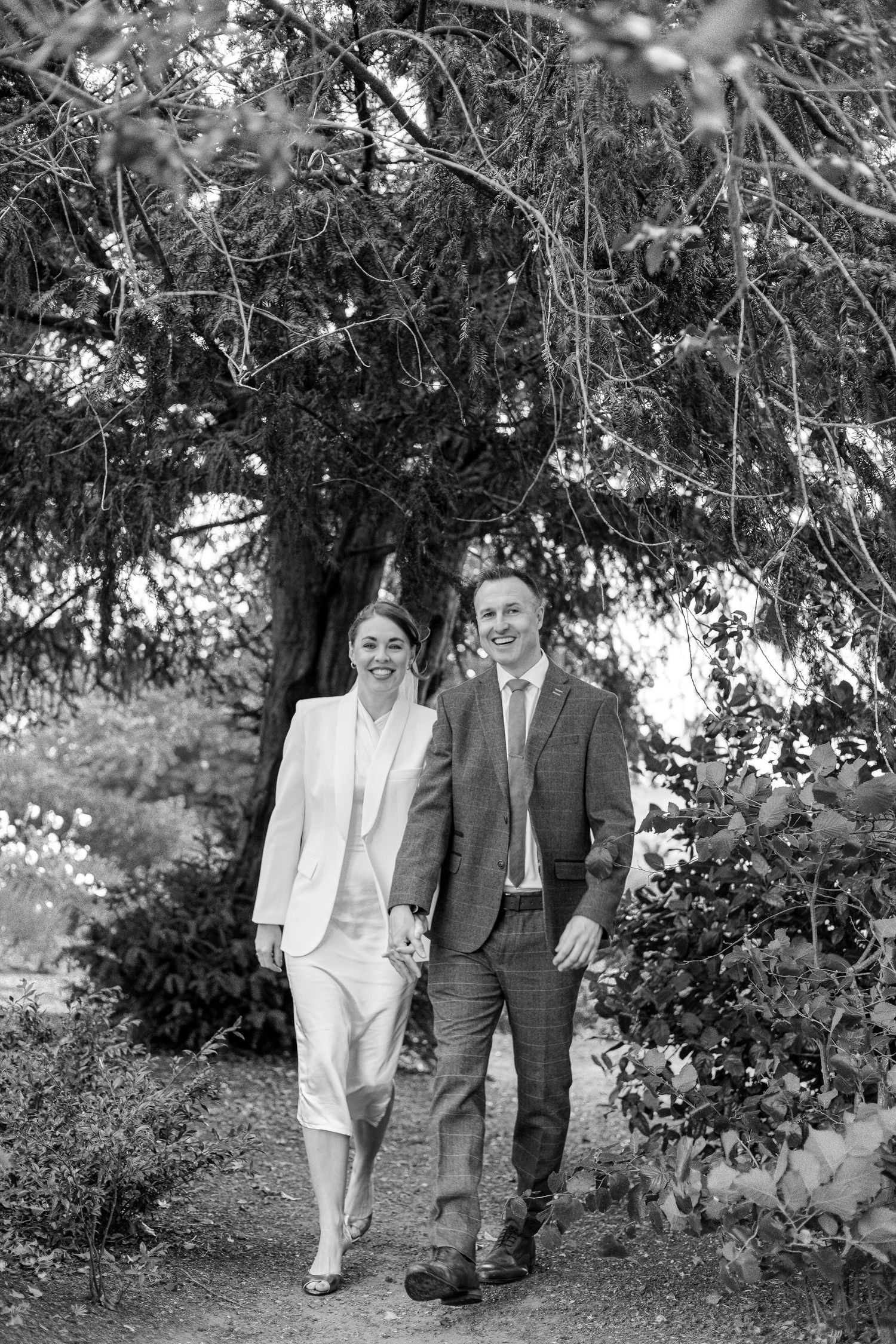 Couple walking together through a garden path under tree branches in black and white.
