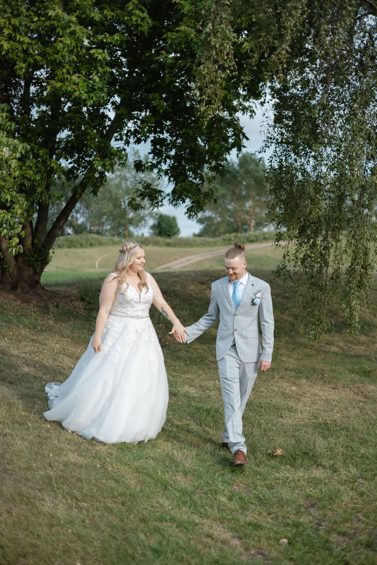 A couple walks hand in hand up a grassy hillside, the bride in white and groom in light gray.