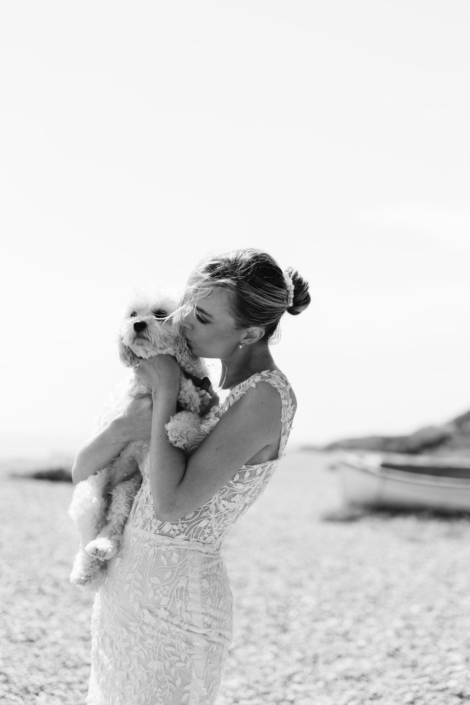 Black and white portraits of a bride holding and kissing a small white dog on the beach.