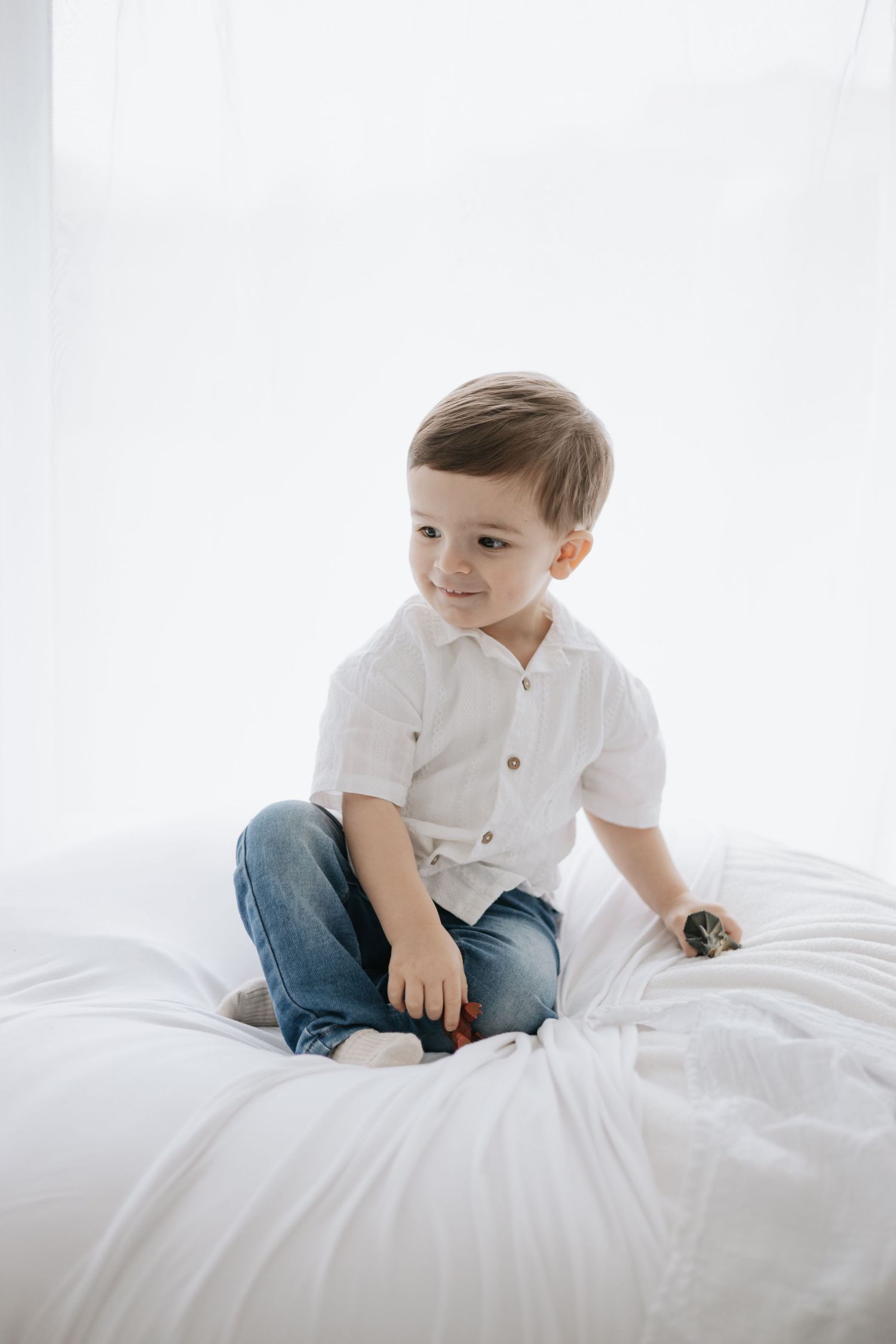Child in white shirt and jeans sitting on white bedding in bright room.