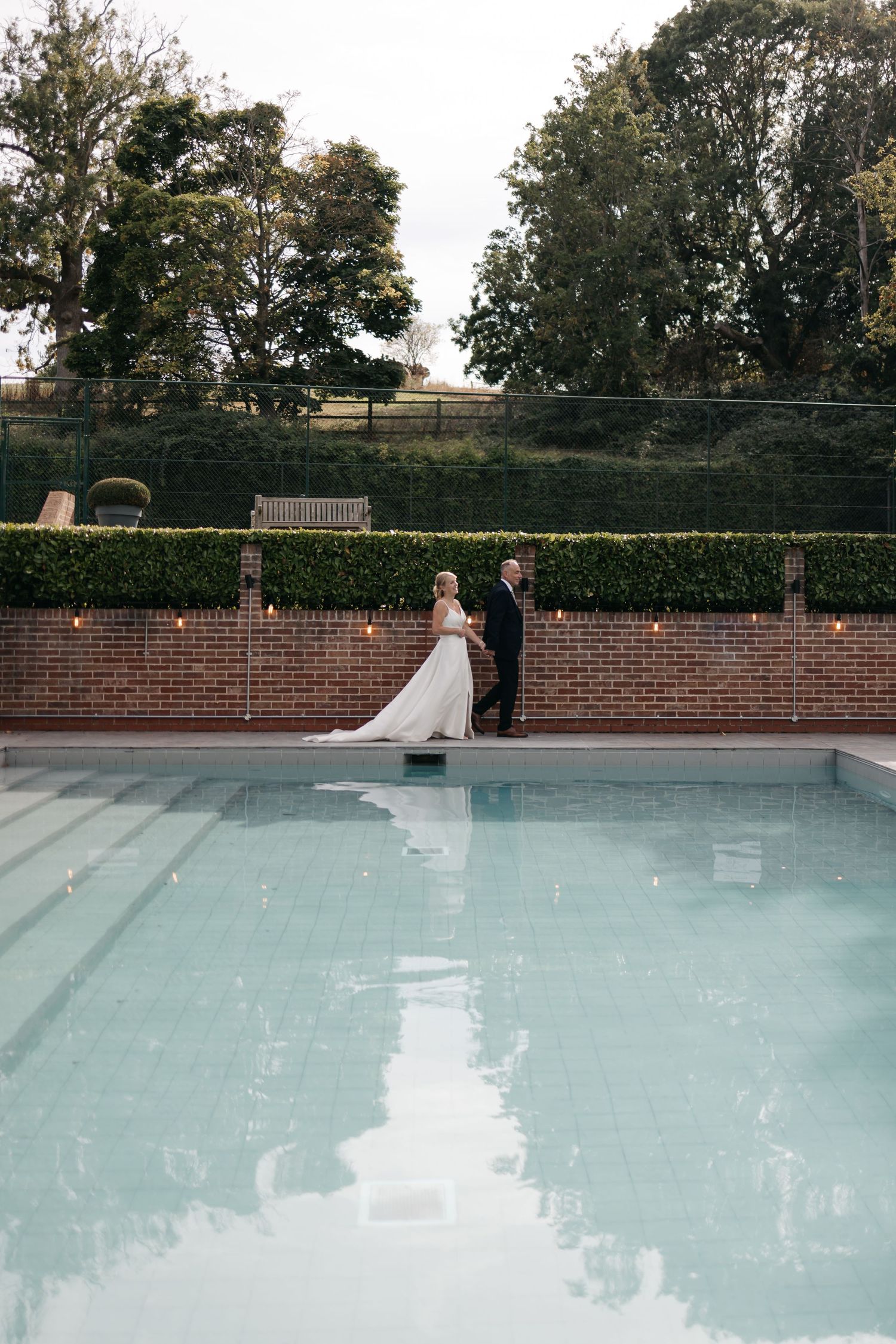 A couple stands beside a pool during their wedding with brick wall and trees in background.