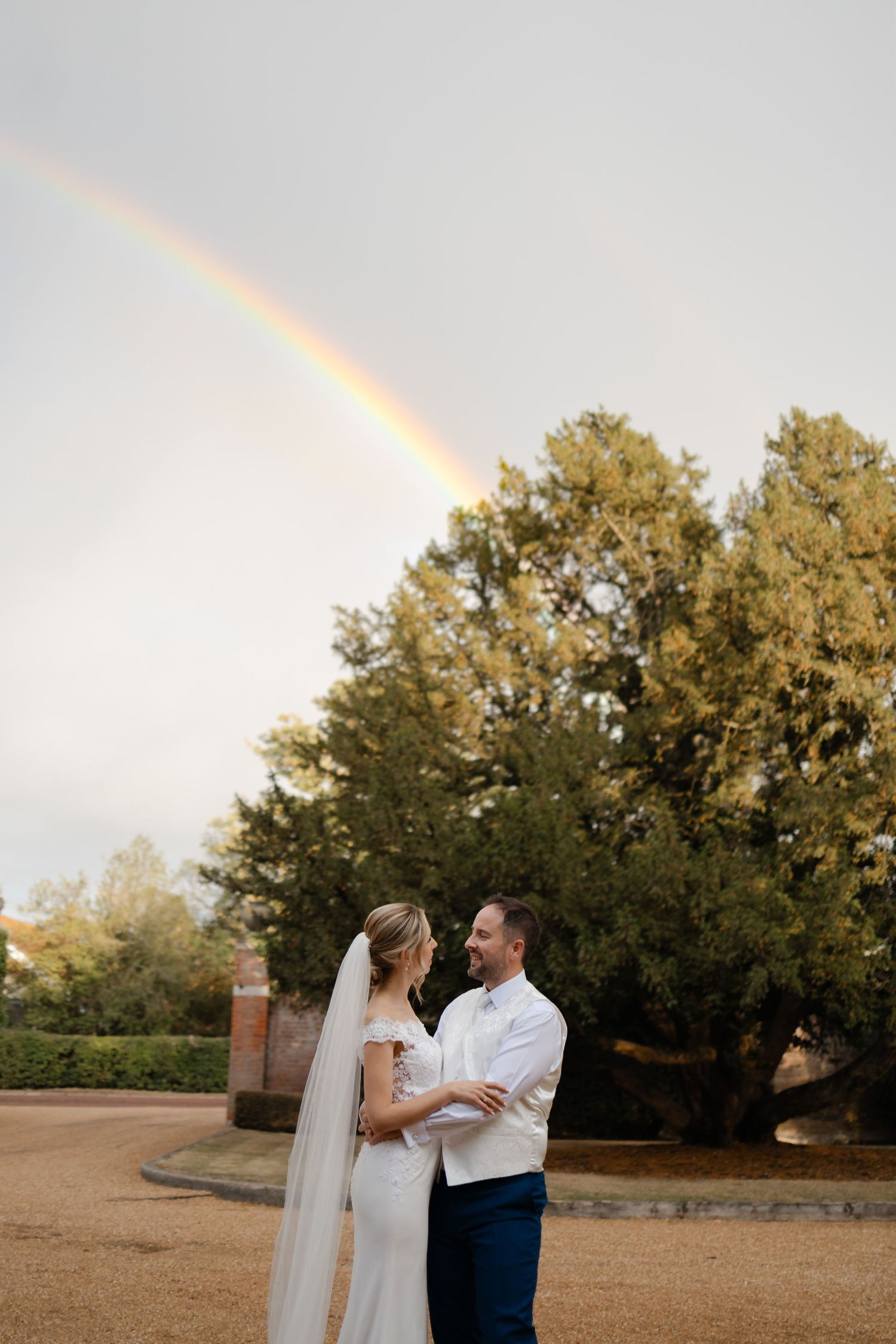 Wedding couple embracing in outdoor setting with rainbow appearing in background.