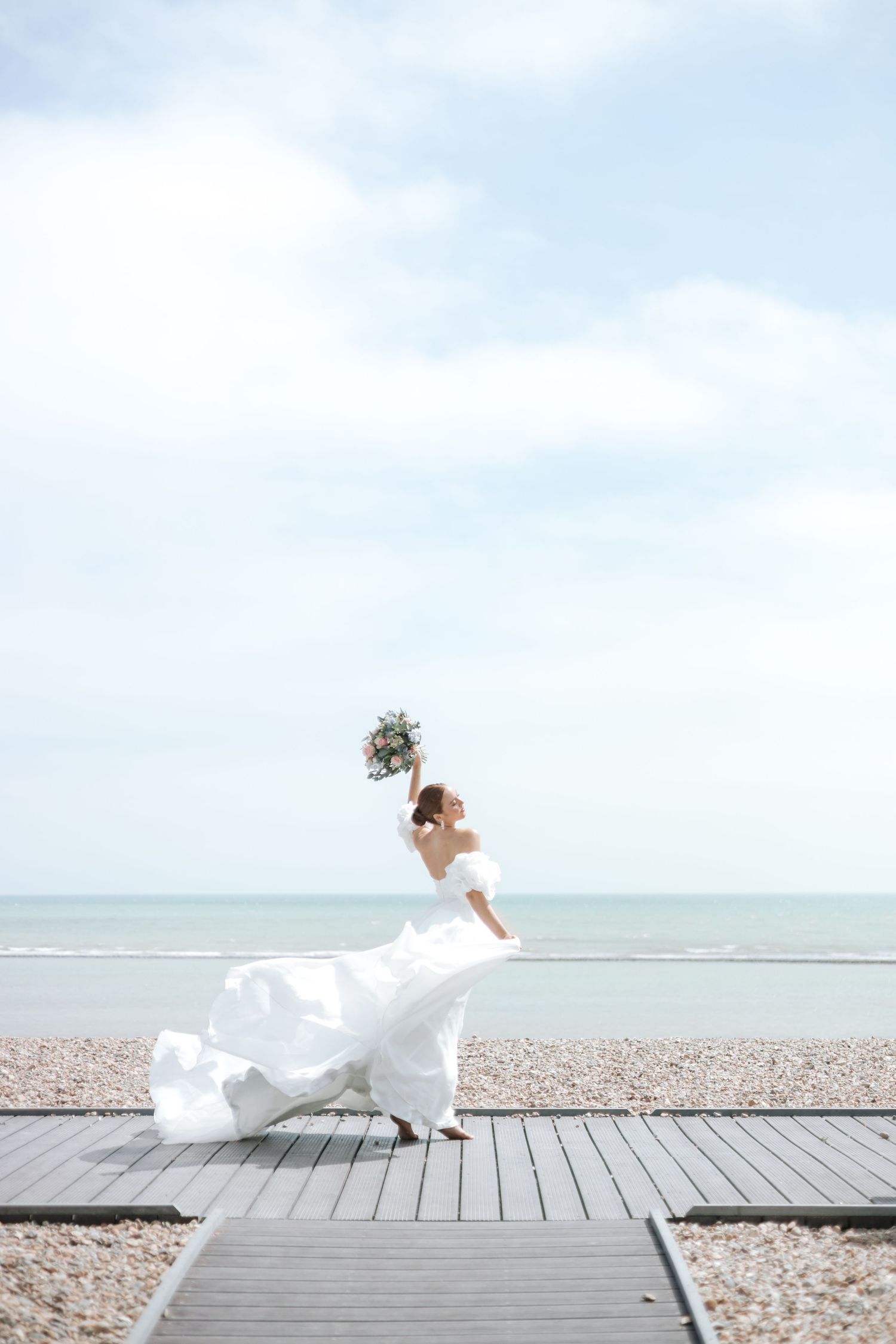 Bride in flowing white dress poses dramatically on wooden boardwalk by the beach.